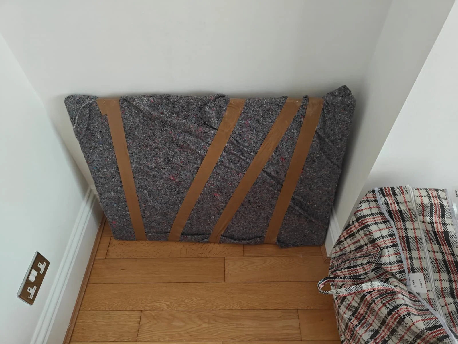 A cushioned piece of furniture or mattress leaning against a wall in a corner, with brown straps securing it, next to a patterned fabric on the floor.