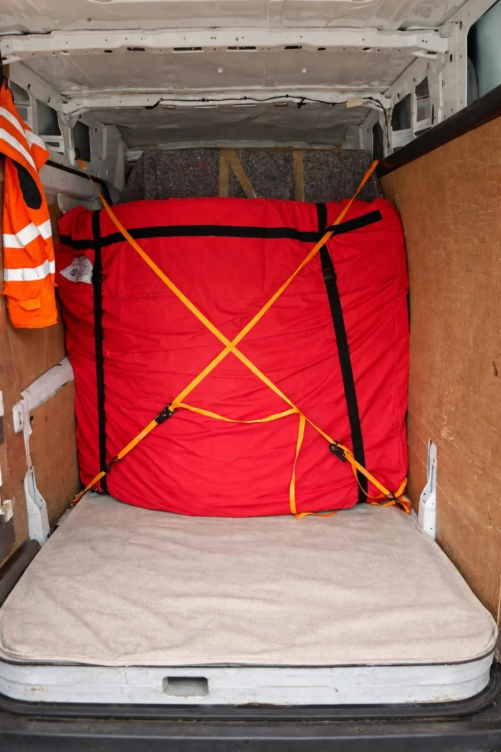 Inside a vehicle cargo area with a red bag secured by yellow straps, a beige mattress on the floor, an orange safety vest hanging on the left, and wooden panels lining the sides.