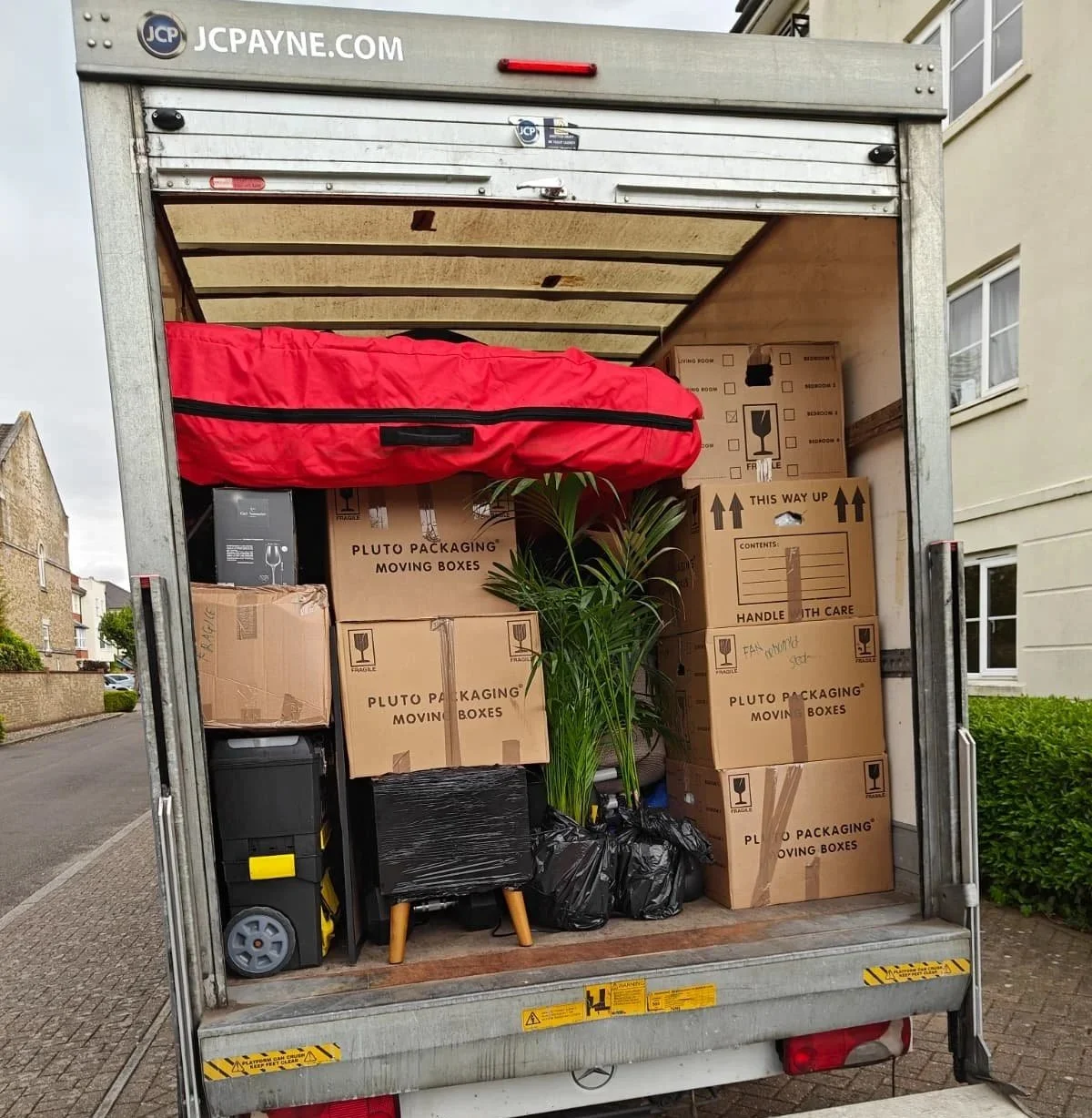 Open moving truck filled with packed boxes, a red bag, a large plant, and some black garbage bags, parked on a residential street.
