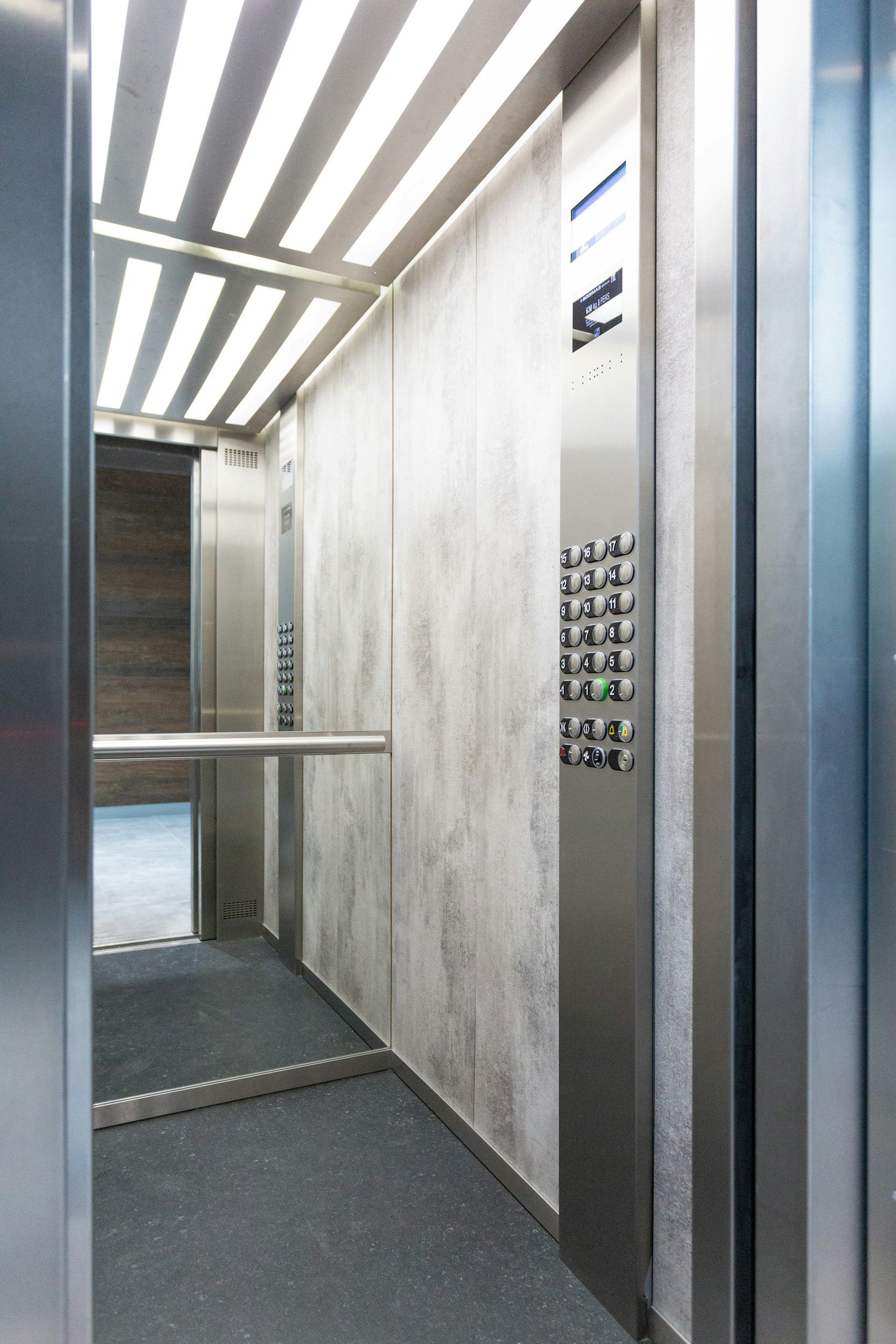 Inside an elevator with stainless steel walls, a control panel with numbered buttons, a digital display, and ceiling lights.