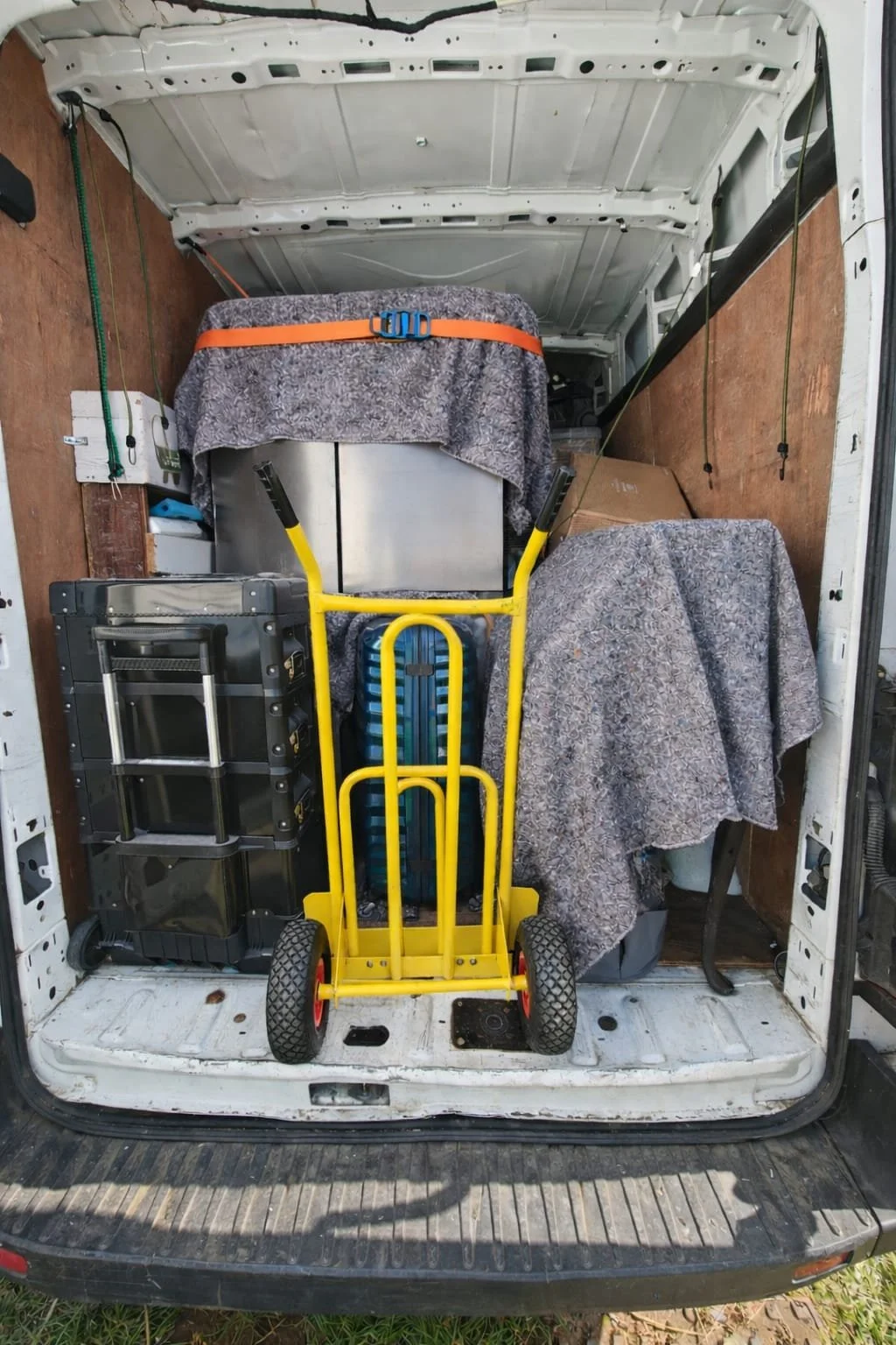 The back of a moving or storage van filled with various household items, including a yellow hand truck, stacked plastic storage containers, boxes covered with patterned blankets, and a cardboard box. The van's interior walls are wooden, and the floor
