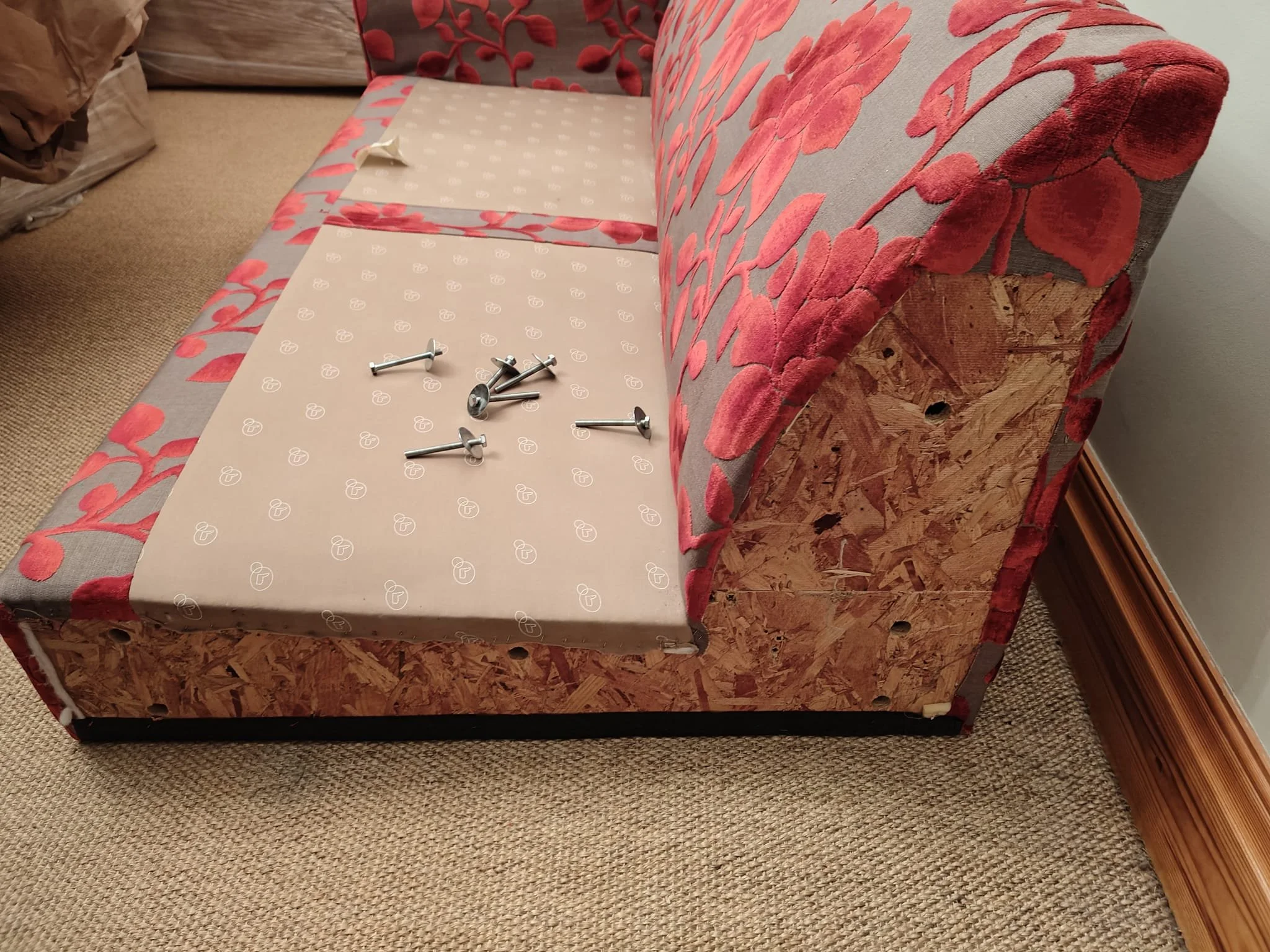 A partially renovated sofa with a cardboard and screws on the cushion, showing a mix of floral fabric and exposed wooden paneling.