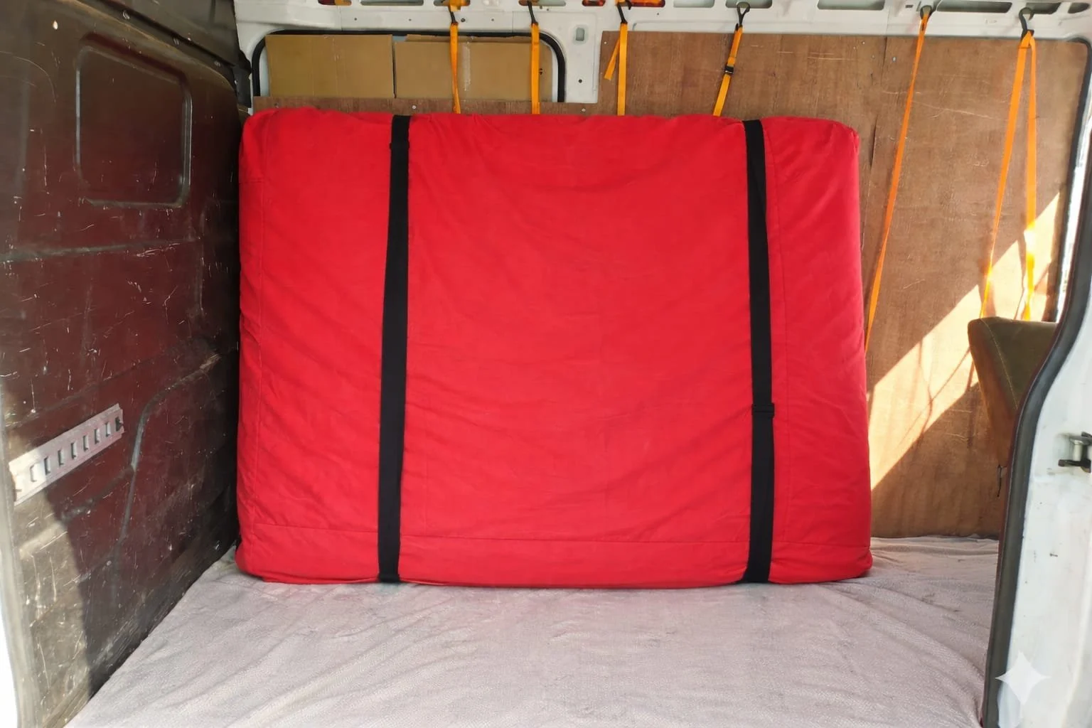 Inside a vehicle with a red mattress secured with black straps, wooden walls, and orange straps on the ceiling for cargo security.