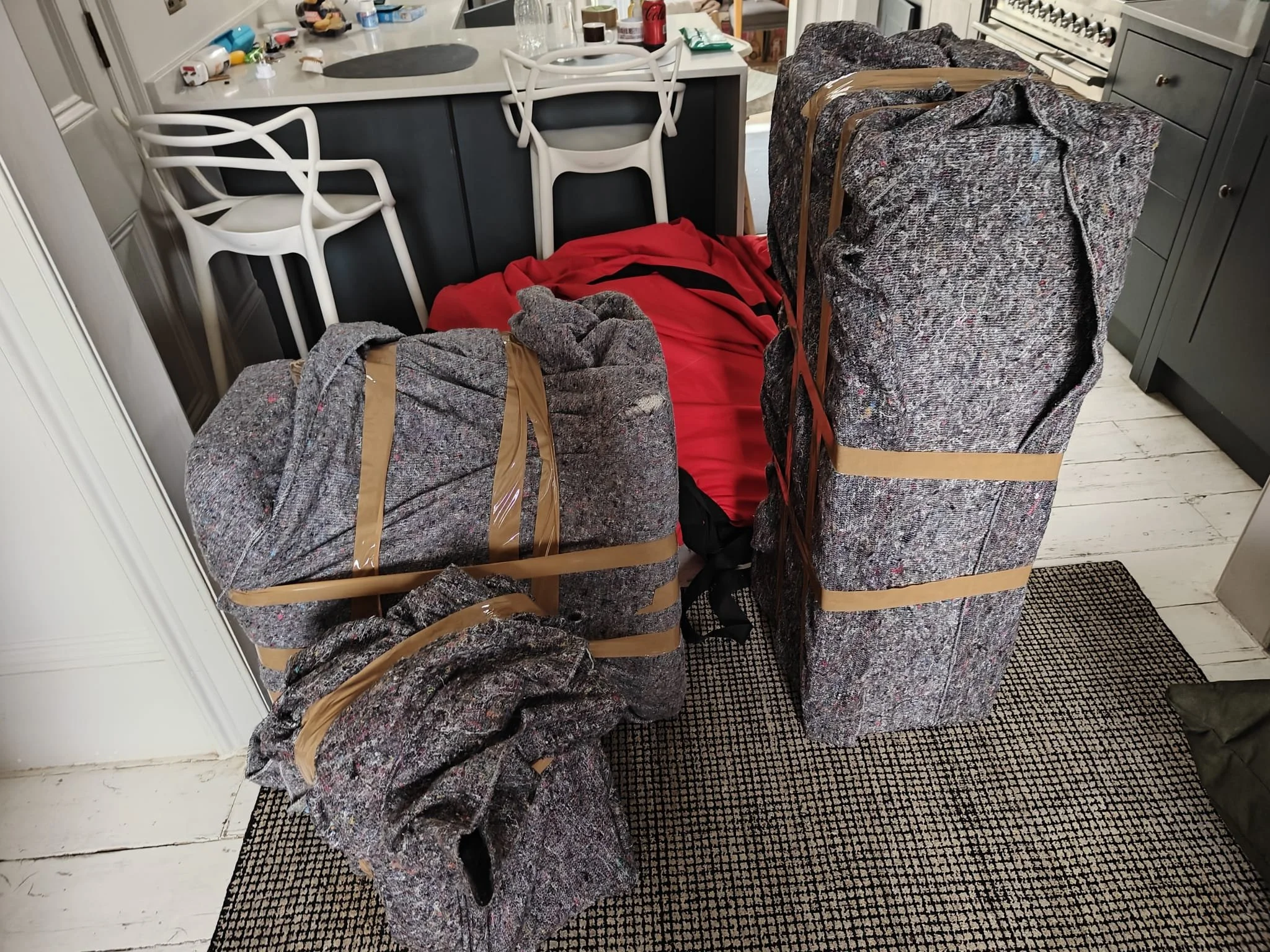 Clothes covered in plastic wrap and tape stacked in a kitchen, near a gray cabinet and charcoal-colored chairs.