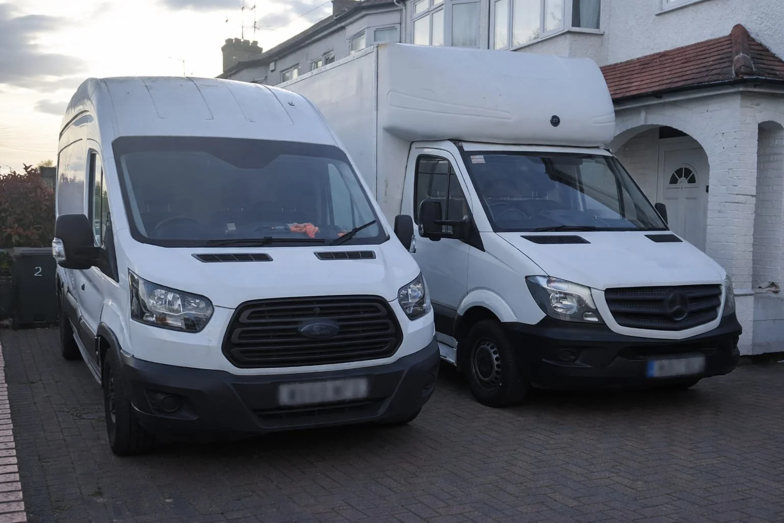 Two white cargo vans parked side by side in a residential driveway, with houses in the background.