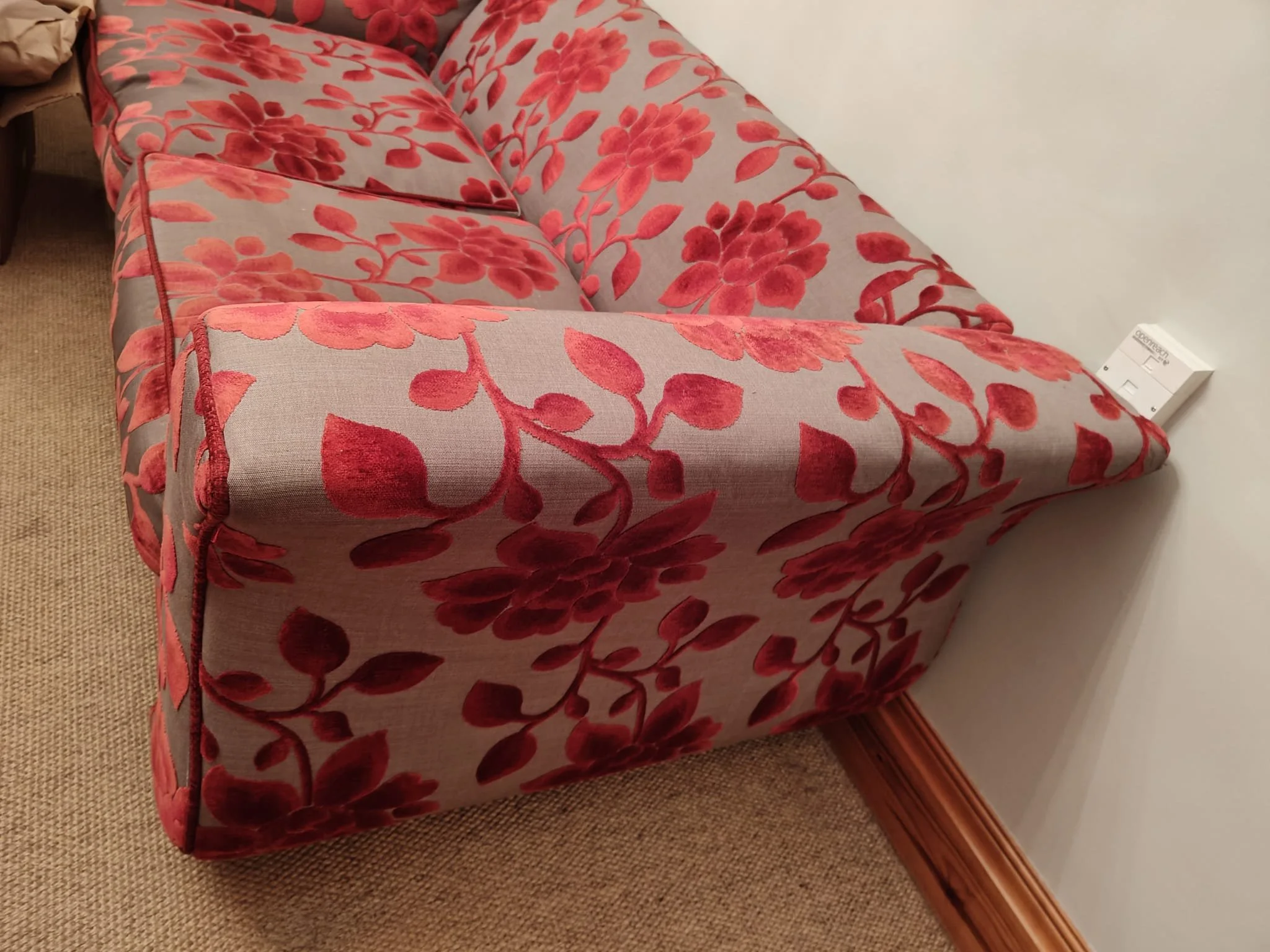 A red upholstered armchair with a floral pattern of large red flowers and leaves, placed on a beige carpet next to a white wall, with a white electronic device plugged into the wall near the chair.