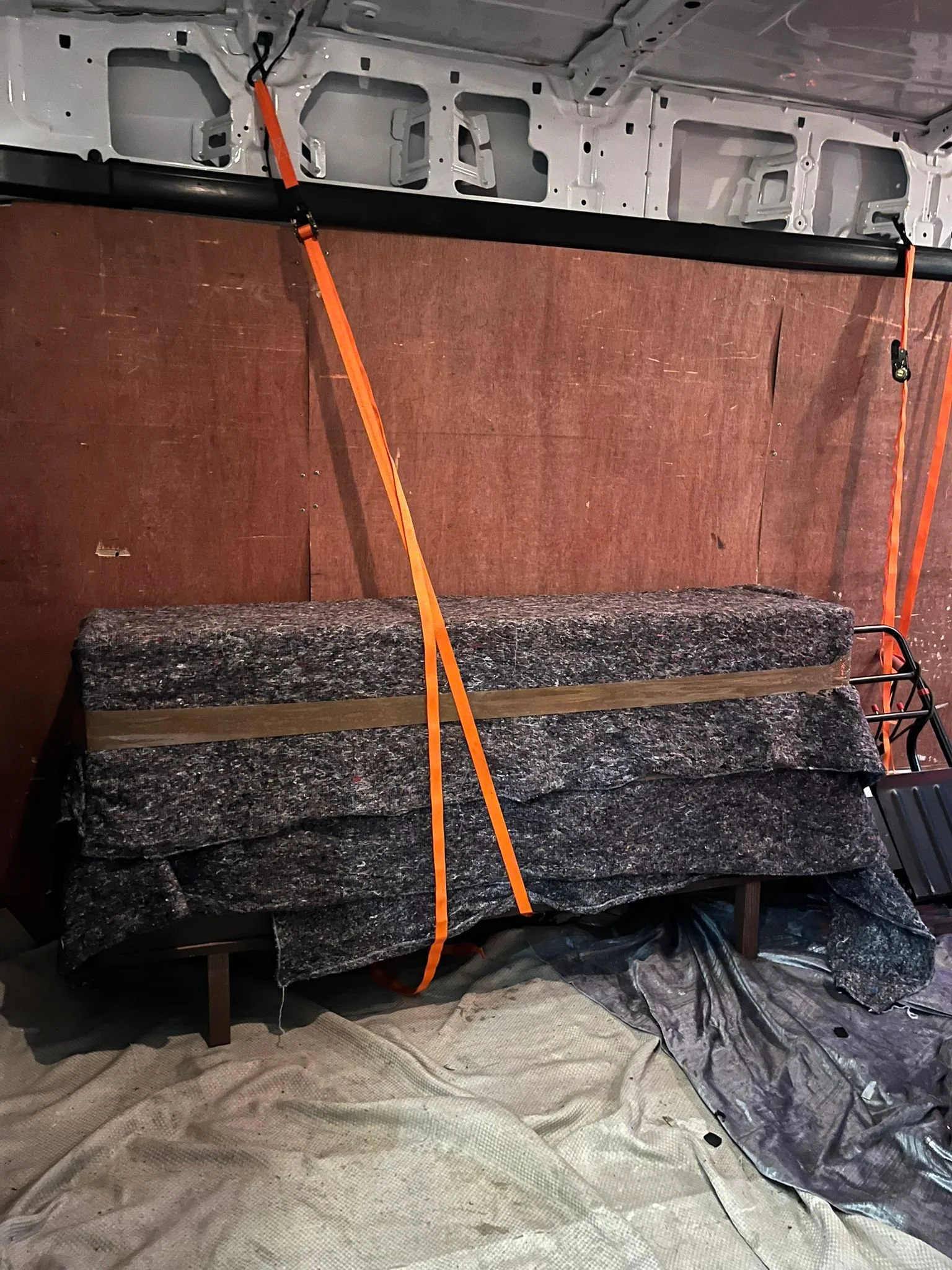 The image shows a room with insulation foam on the floor, a piece of fabric, and a workspace with a wooden panel, a large rock, and orange straps hanging from the ceiling. The ceiling and wall have unfinished drywall and wooden siding.