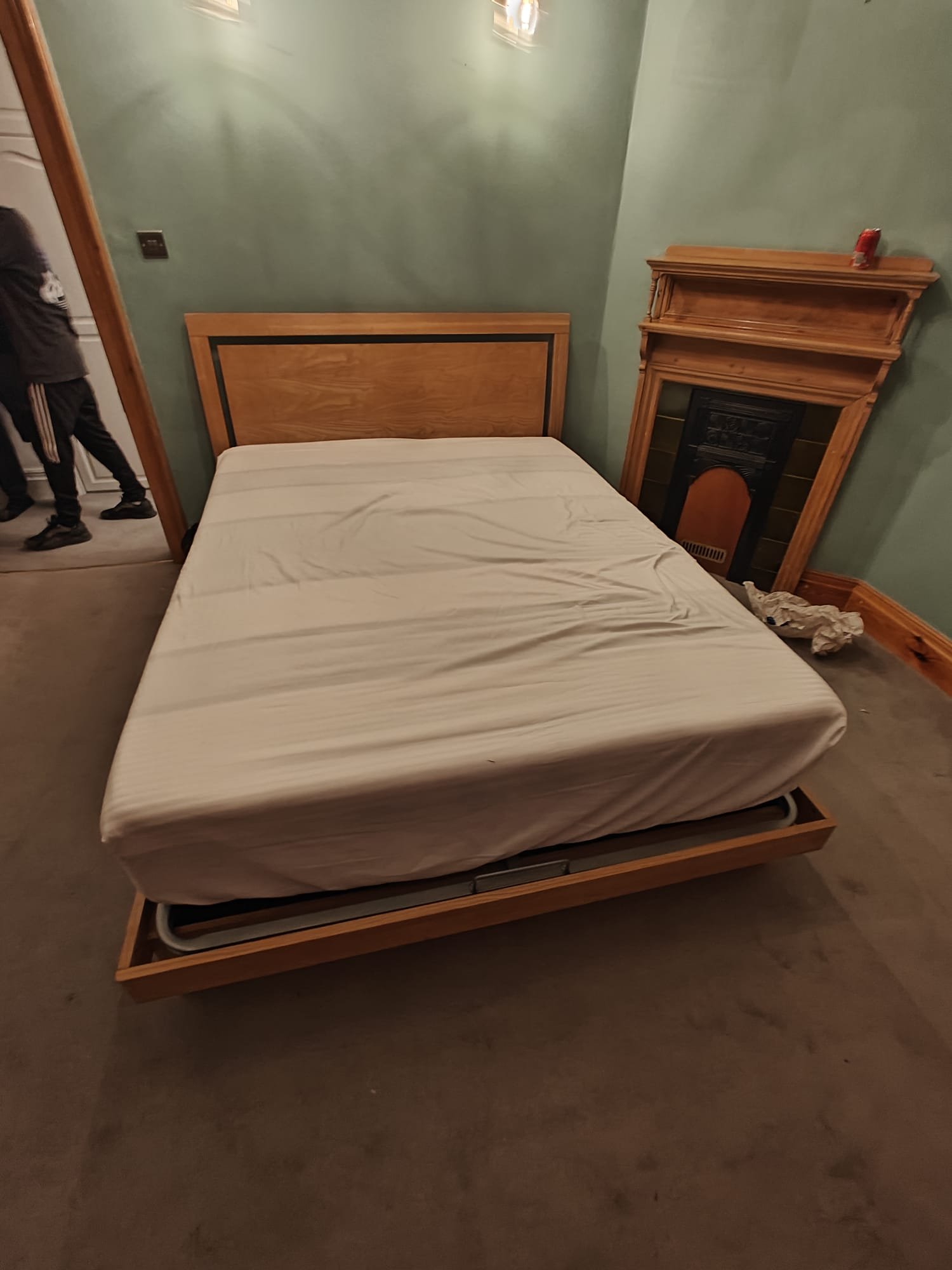 A bed with a wooden headboard and a white mattress in a room with light green walls. To the right of the bed, there is a wooden fireplace mantel with a small object on top. There is a crumpled piece of paper on the floor near the fireplace. On the le