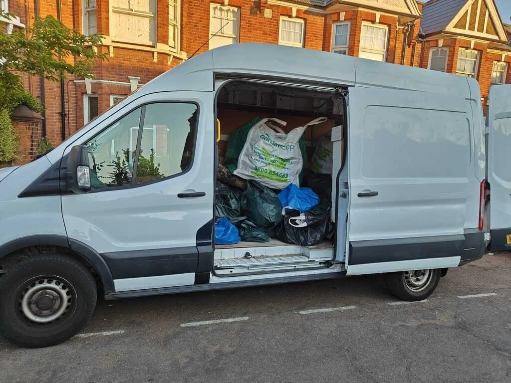 White cargo van with its sliding door open, filled with trash bags and garbage, parked on a residential street with brick houses and trees in the background.