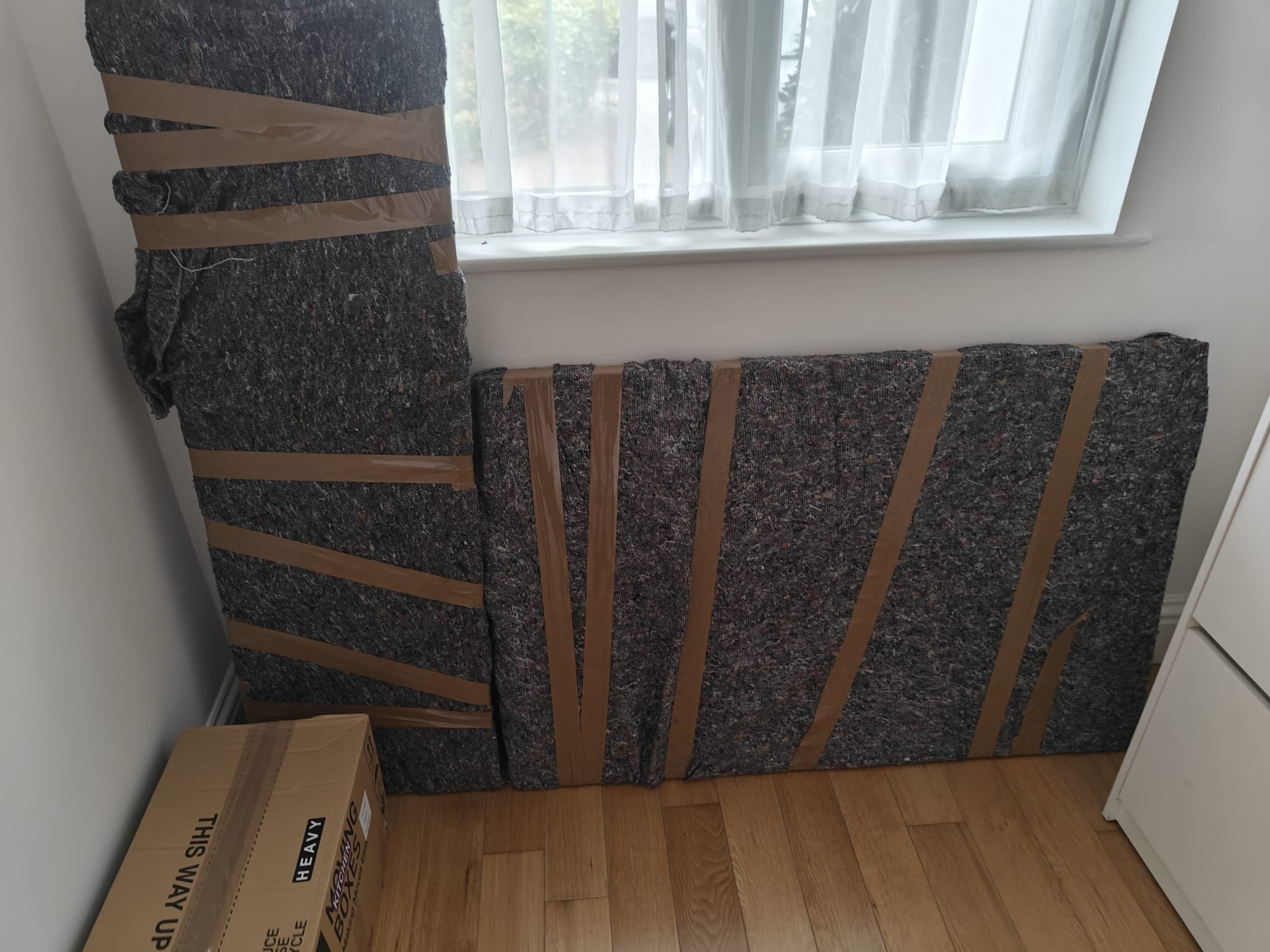 A bedroom corner with a bed frame, no mattress, pinkish-brown tape holding parts together, a cardboard box nearby, and a window with sheer curtains