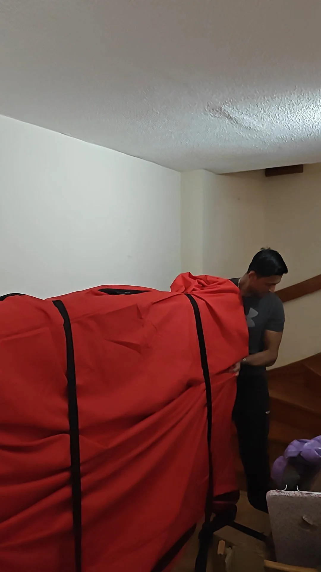 A man in a gray shirt is standing and bending over next to a large red item or furniture, in a room with wooden stairs and white walls.