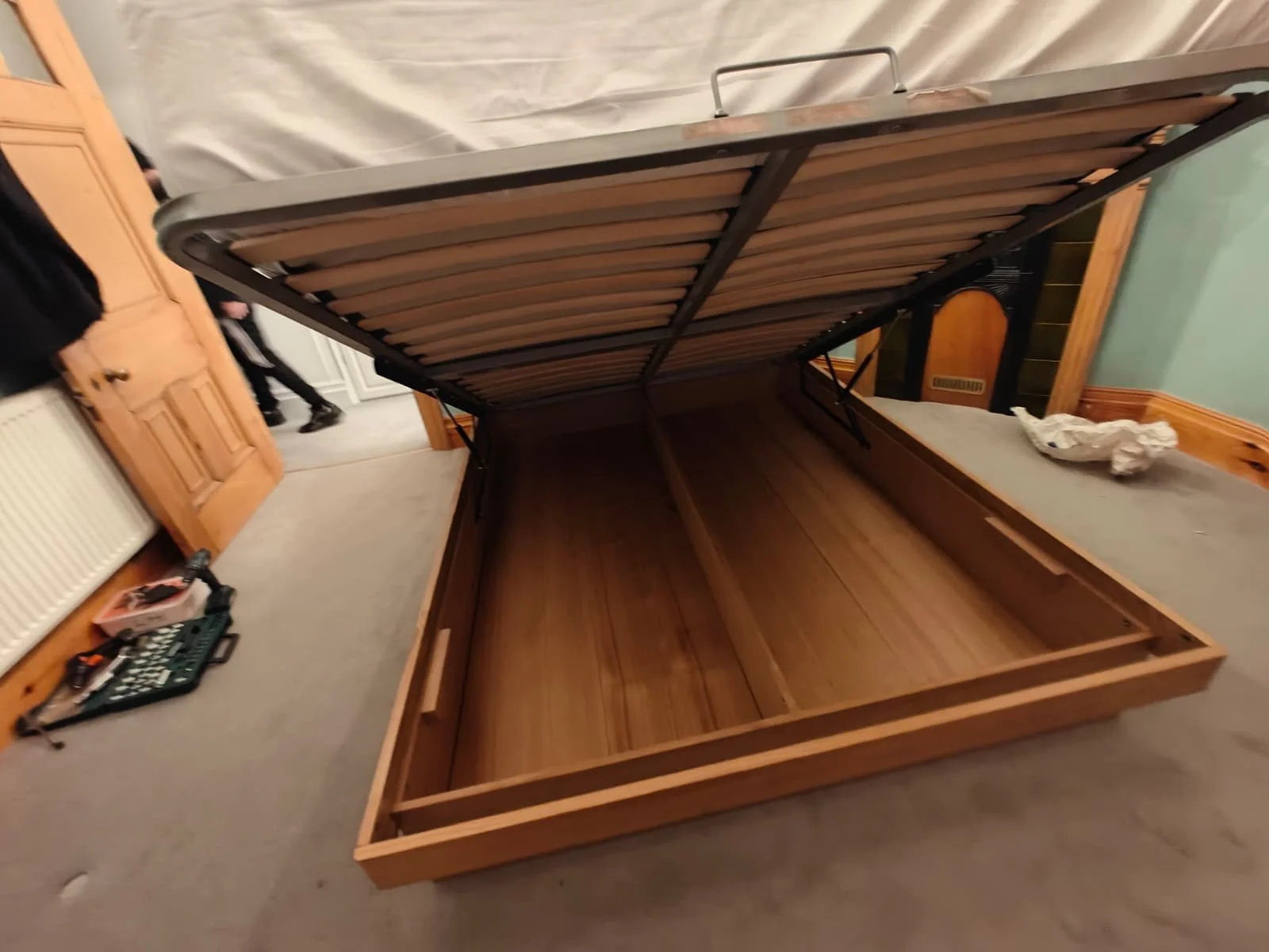 Unassembled wooden bed frame with a mattress stacked on top in a bedroom. The bed frame is open and empty, showing the slats. There are various tools on a nearby table and a person standing in the background.
