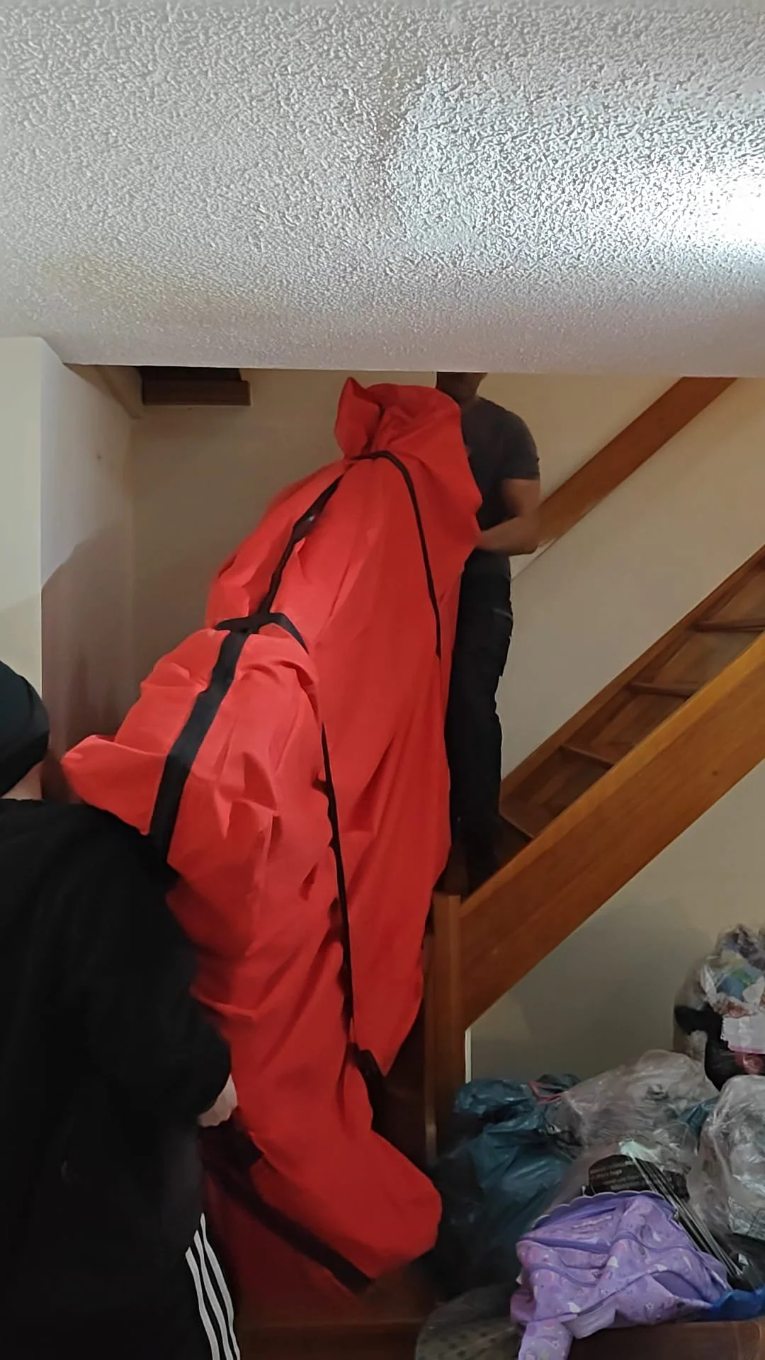 People moving or storing belongings in a room under a staircase, with bags and items on the floor.