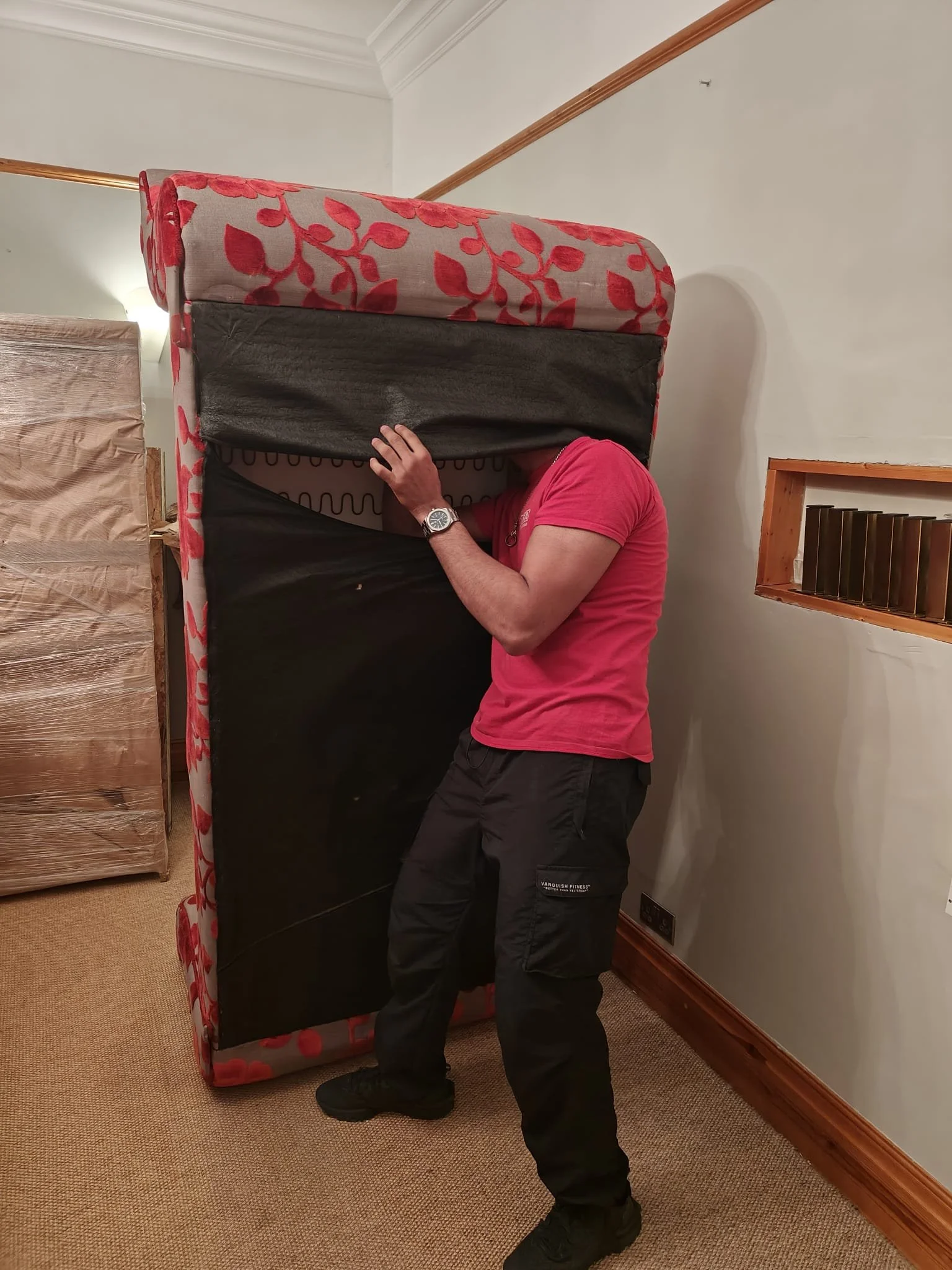 A person with a red shirt is lifting a mattress on a black bed frame inside a room with beige carpet and white walls.