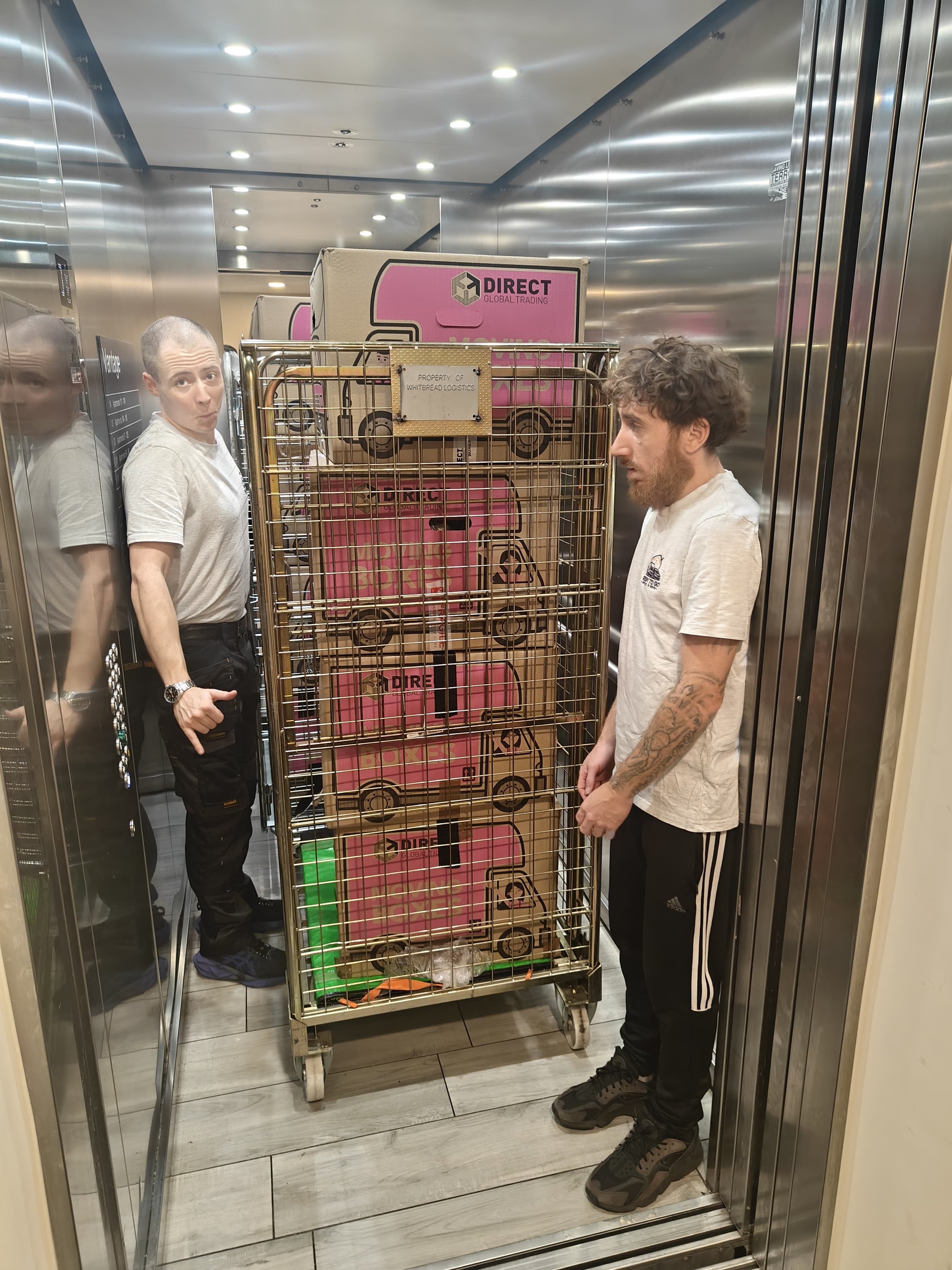 2 movers from Ready To Go House Movers And Removals inside a lift with their roll cage cart filled with boxes and going down into their Luton van for loading. During a flat move in West London.