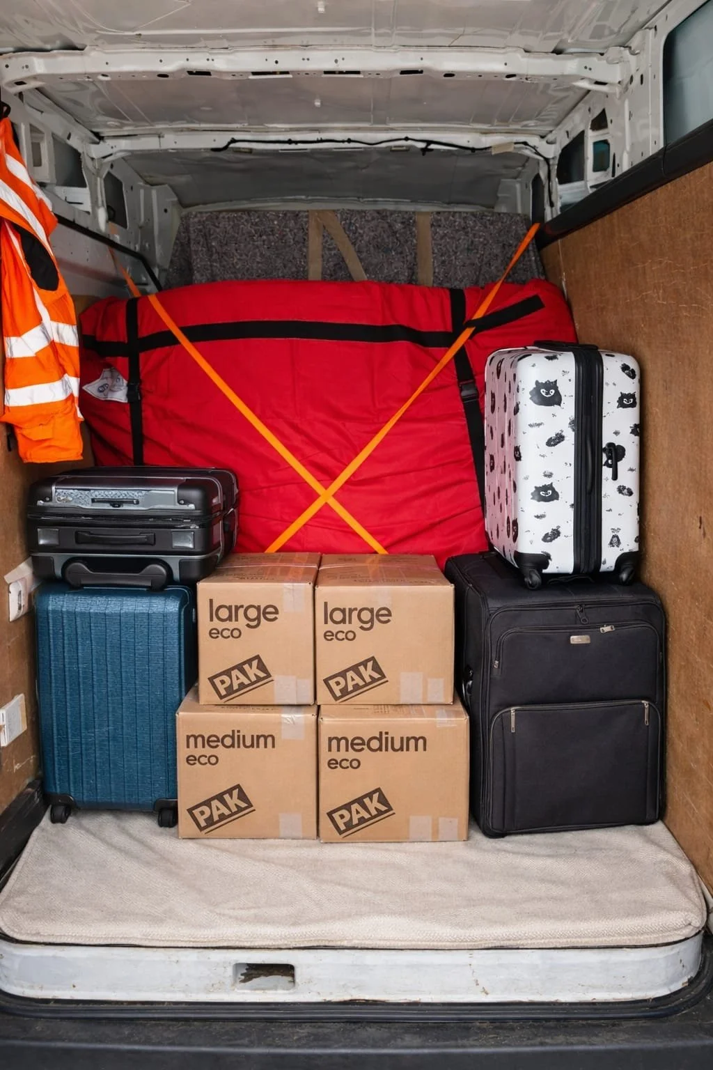 The back of a vehicle packed with luggage, boxes, and a red soft-sided suitcase secured with orange straps, along with an orange safety vest hanging in the corner.
