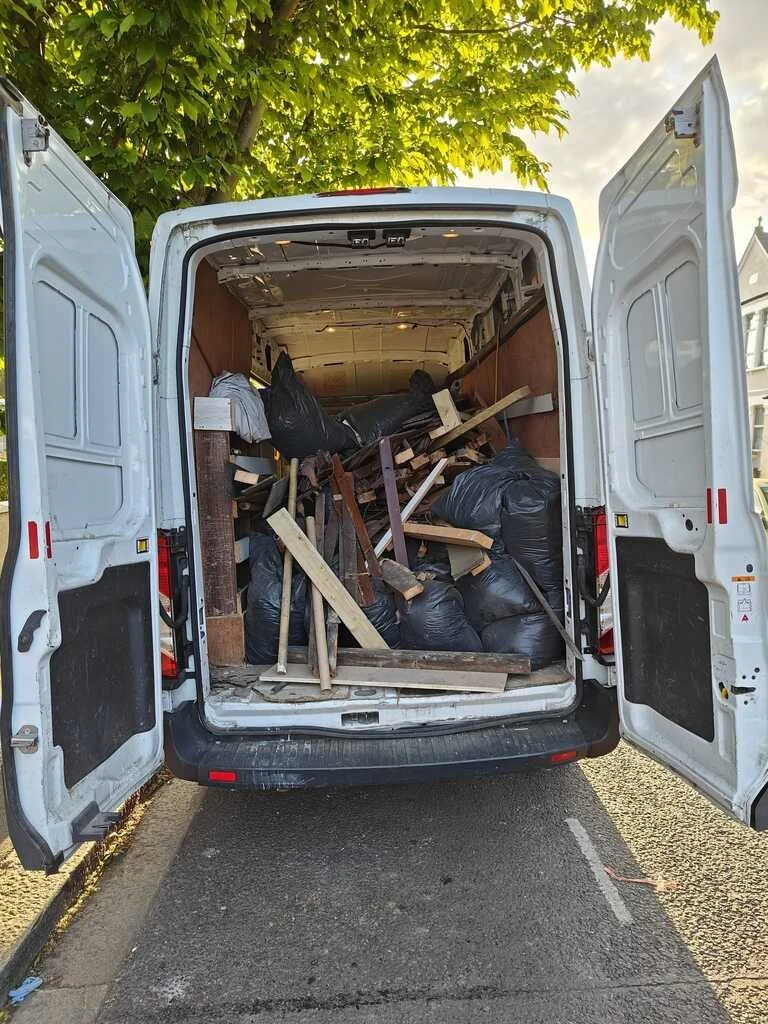 Open van filled with black garbage bags and assorted wooden debris, with a tree and houses in the background.