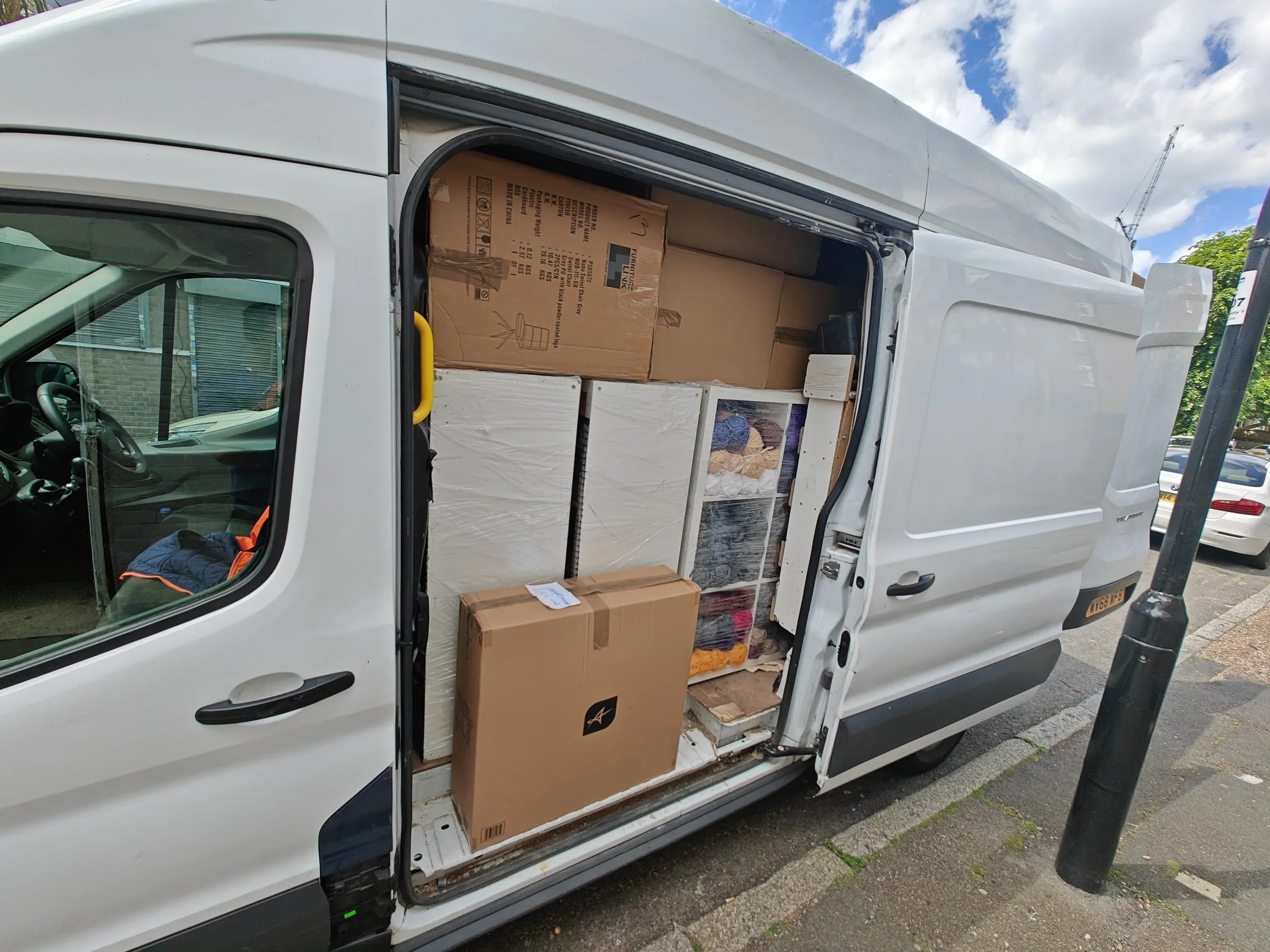 Open side door of a white delivery van filled with boxes and packages. The van is parked outside during daytime with a blue sky and some clouds visible.