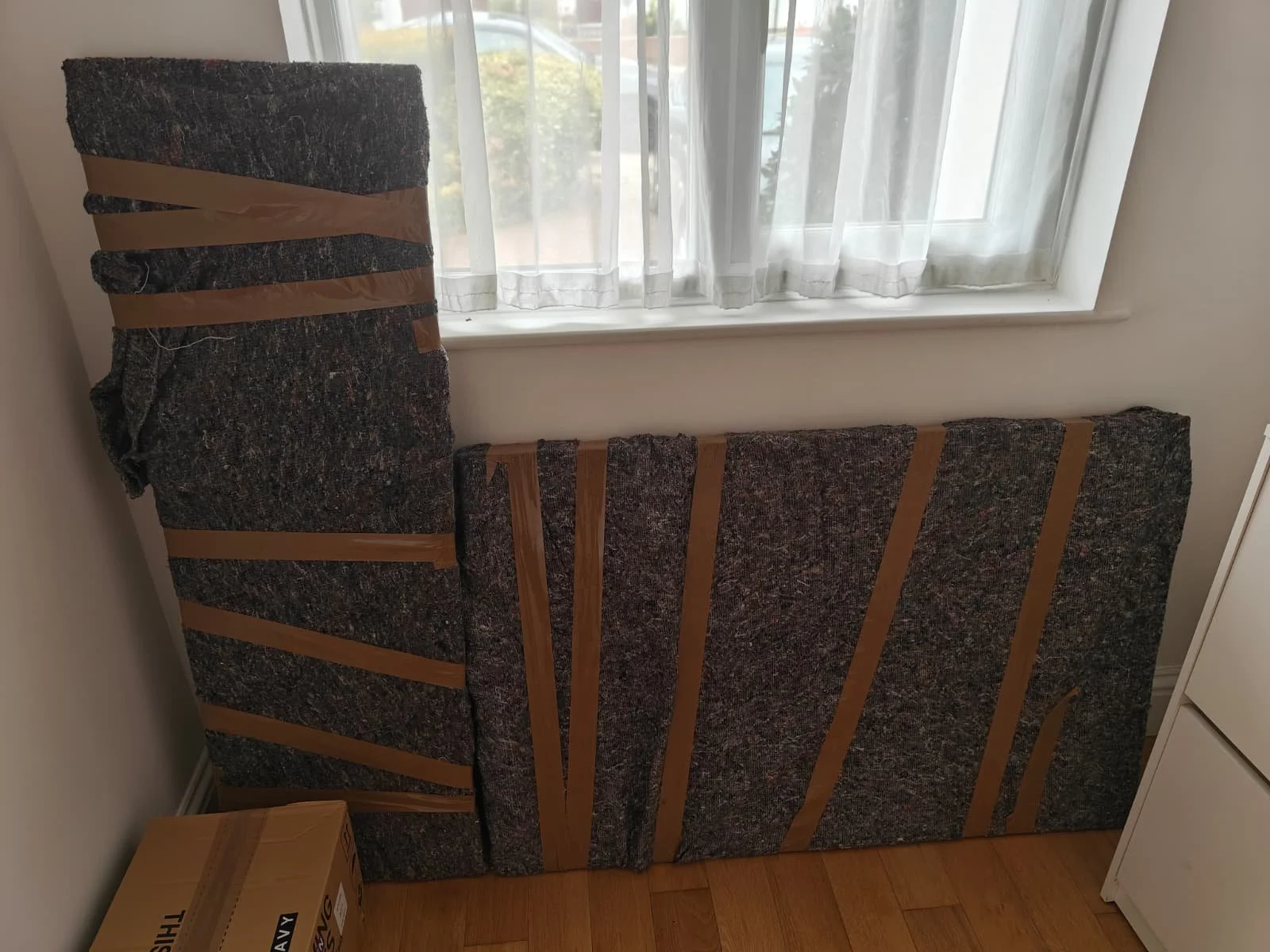 View of two large, wrapped pieces of furniture or panels with striped fabric and tape, leaning against a wall near a window with sheer curtains.