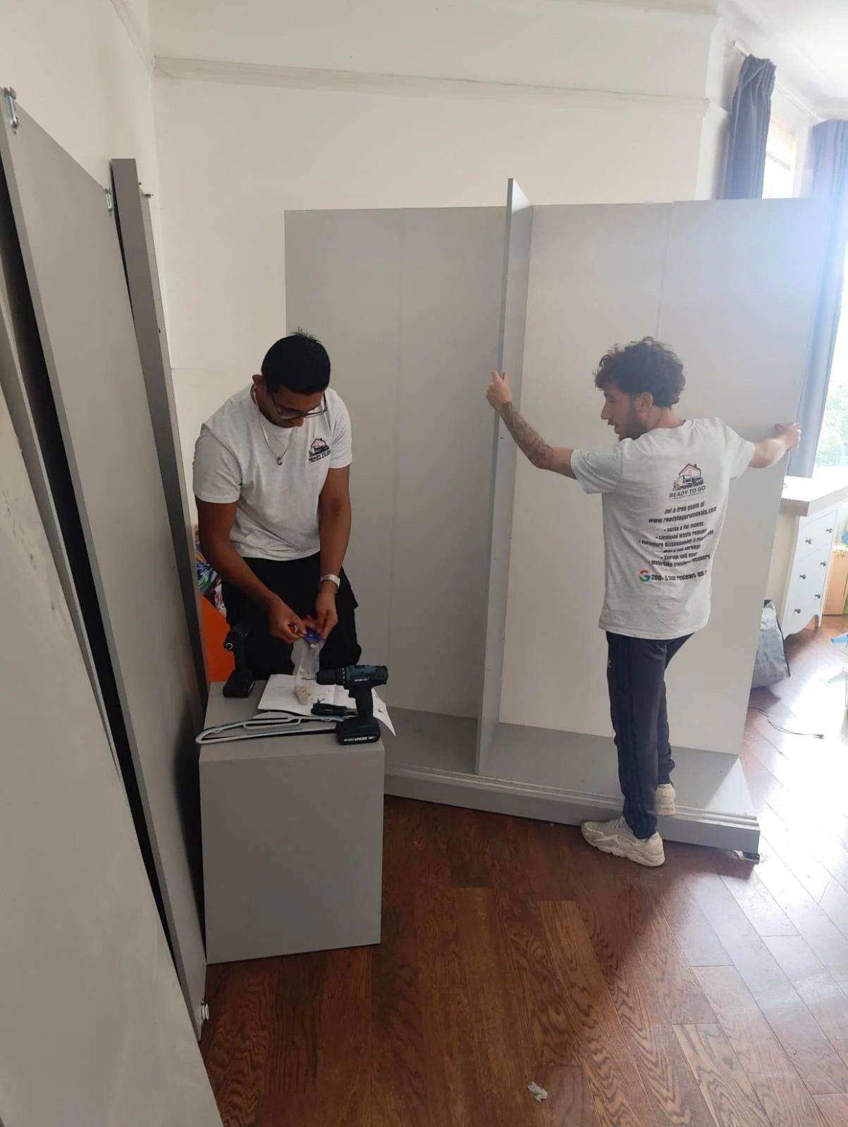 2 removal men from ready to go house movers and removals working on taking apart a large grey wardrobe
