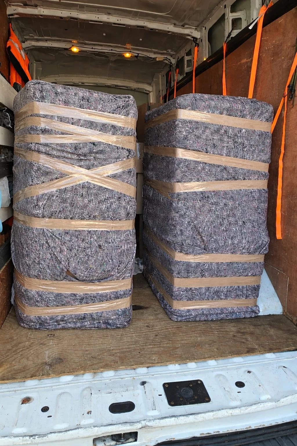 Two large bundles of bluish-gray felt wrapped with wooden slats and strapped with tan packing tape, placed inside the back of a cargo van with wooden interior walls.