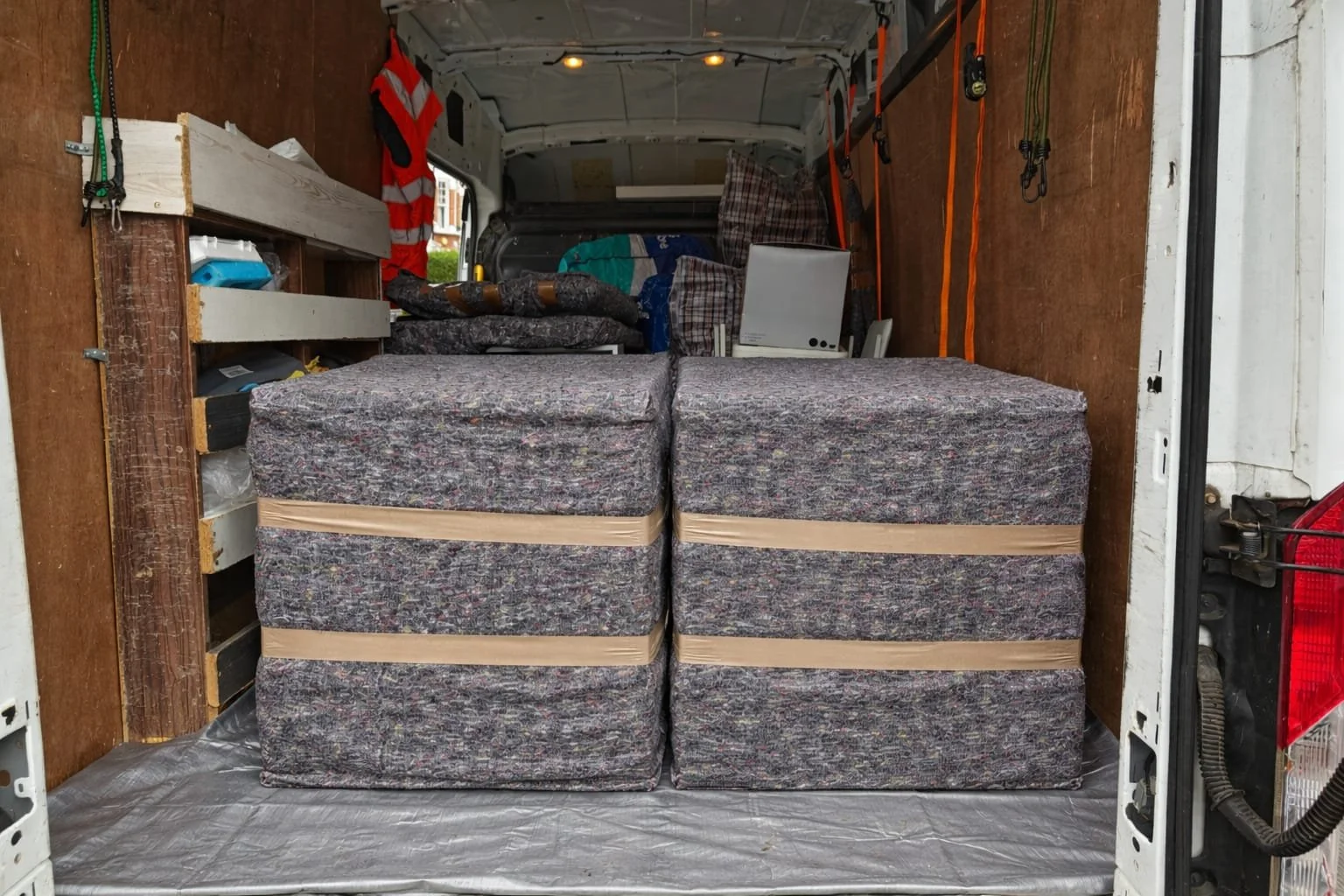Inside a moving van with stacked used mattresses at the entrance, shelves with various items, and secured with straps.