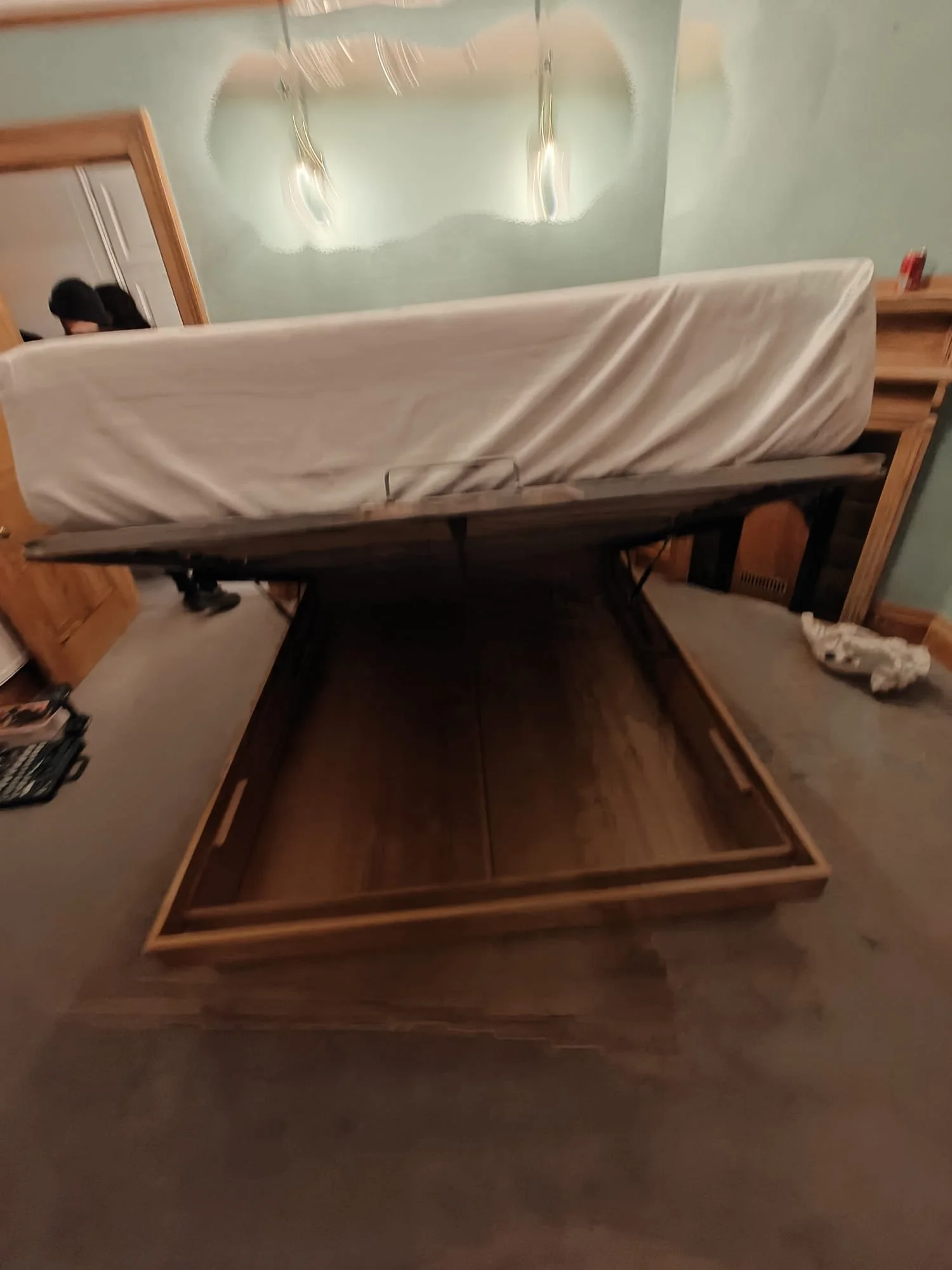 A bed with a wooden frame and a mattress, partly raised, in a room with a green wall and a mirror. There is a side table with a red object on it, and a piece of cloth on the floor.