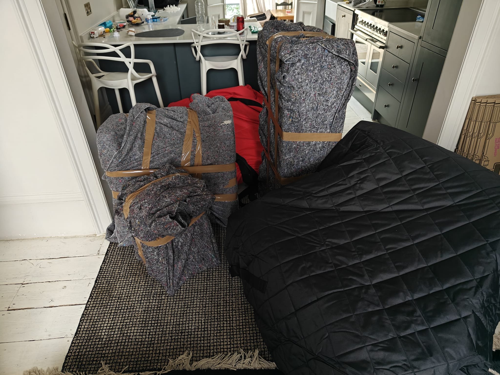 A kitchen with a cluttered countertop and three large, packed bags in the foreground. The kitchen has a stove, oven, and cabinets, with white chairs and a dining table in the background.