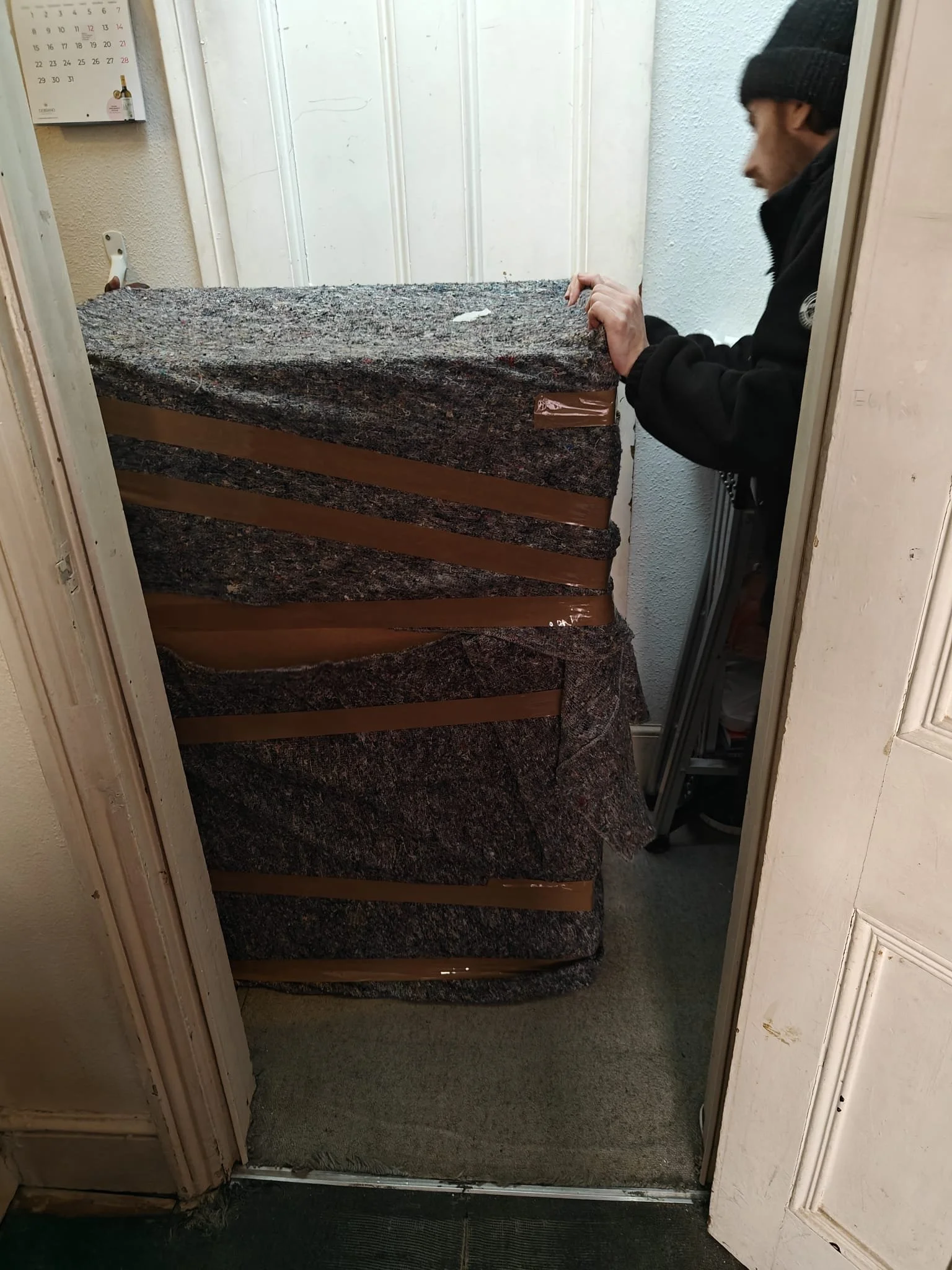 A man with a beanie and black jacket leaning over a stacked pile of granite countertops in a doorway, with a calendar on the wall and a carpeted floor.