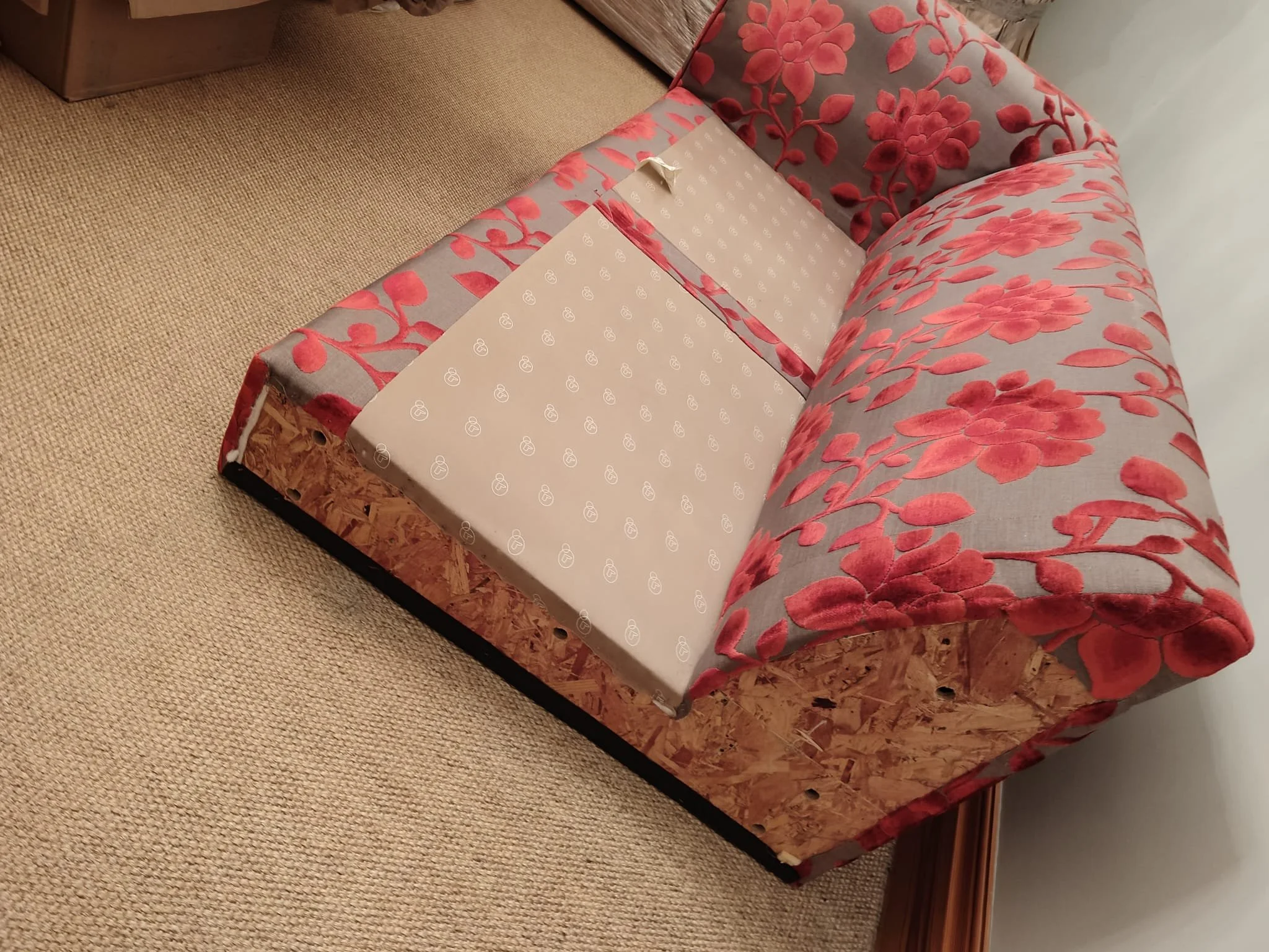 Close-up of a sofa with a floral pattern in shades of gray and red, draped over a wooden base, placed next to a beige carpet and wooden furniture.