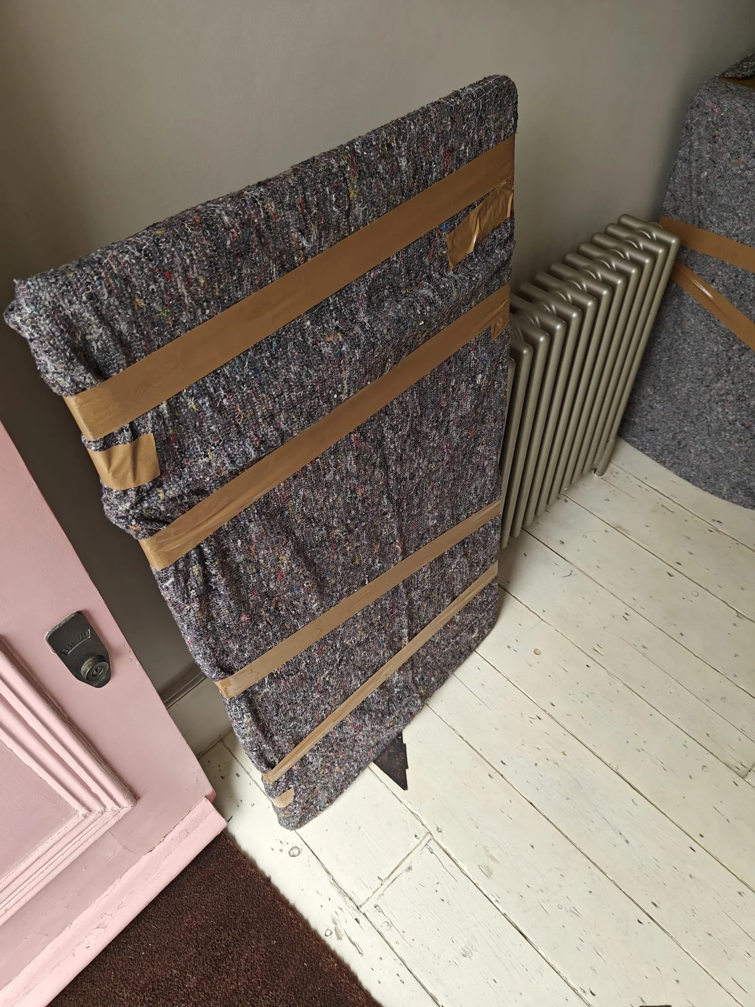 Stack of carpet samples leaning against a wall next to a vintage radiator, with a pink door and white wooden flooring.