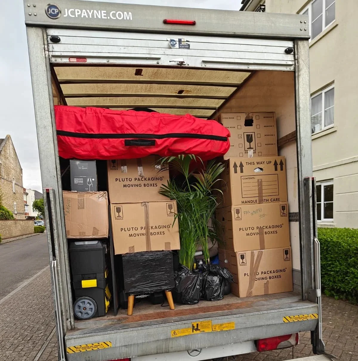 The back of a moving truck filled with packed boxes, a potted plant, a red bag, and a small black furniture piece, with residential houses nearby.