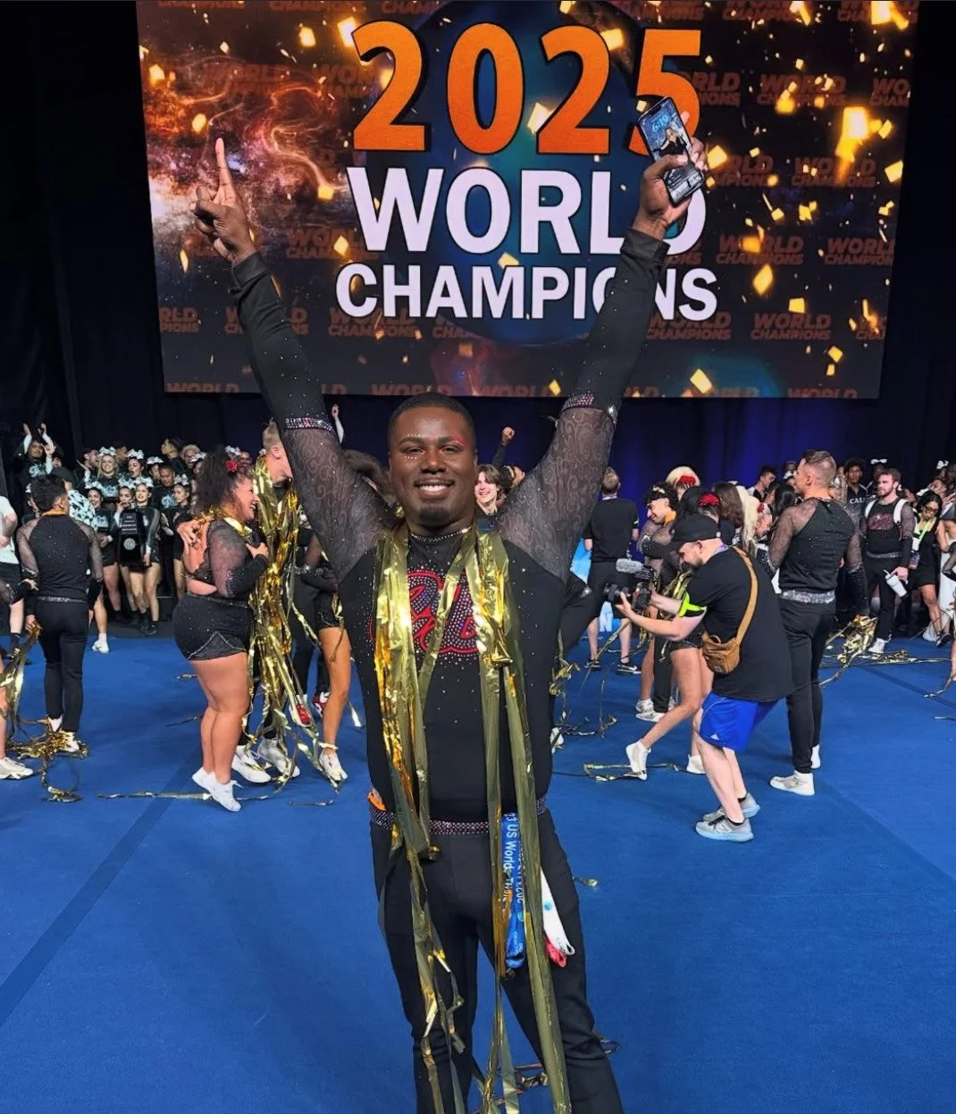 A man celebrating at the 2025 World Cheerleading Championships, with arms raised, surrounded by cheerleaders and spectators, in front of a large screen displaying the event name and year.
