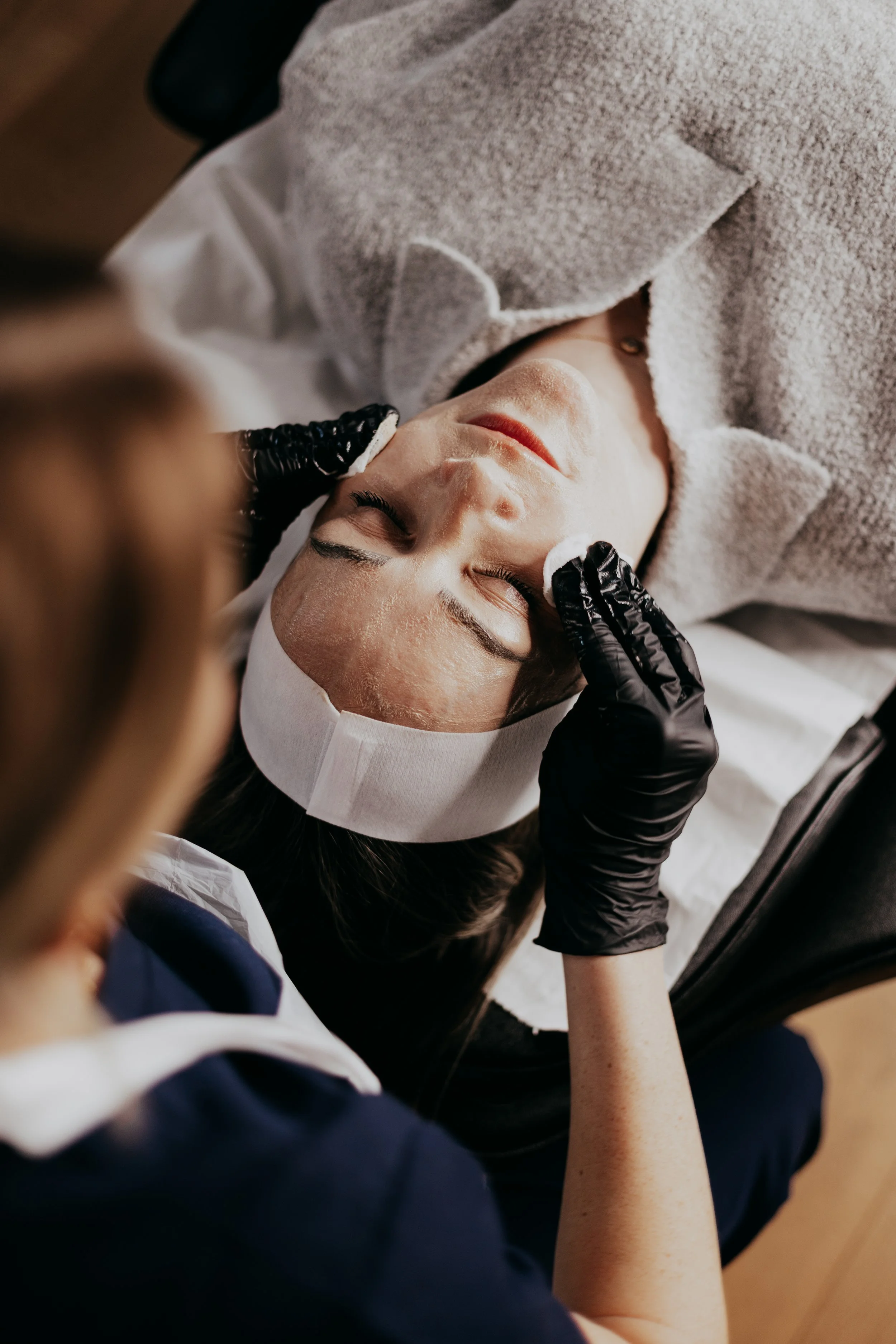 Medical or skincare treatment setup on a black cart, including bottles, hand sanitizer, towel, face mask, headset, and miscellaneous items, with a mirror and office space with a chair and laptop in the background.