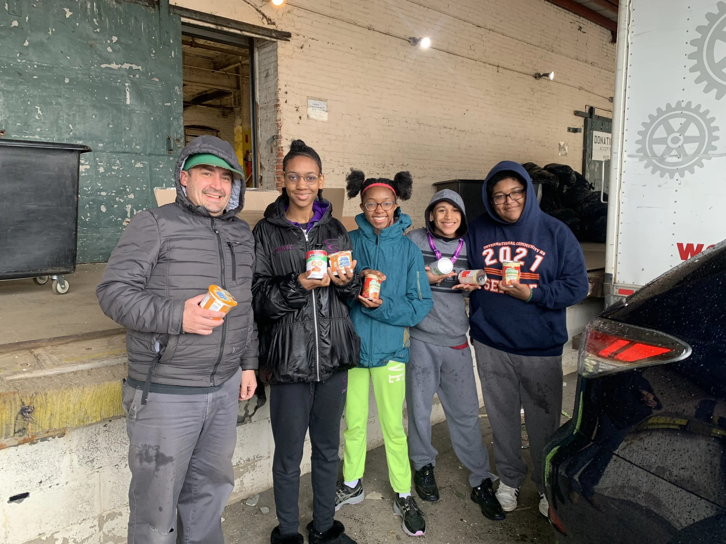 Five people standing outdoors in front of a building, holding canned food. They appear to be participating in a food donation event.