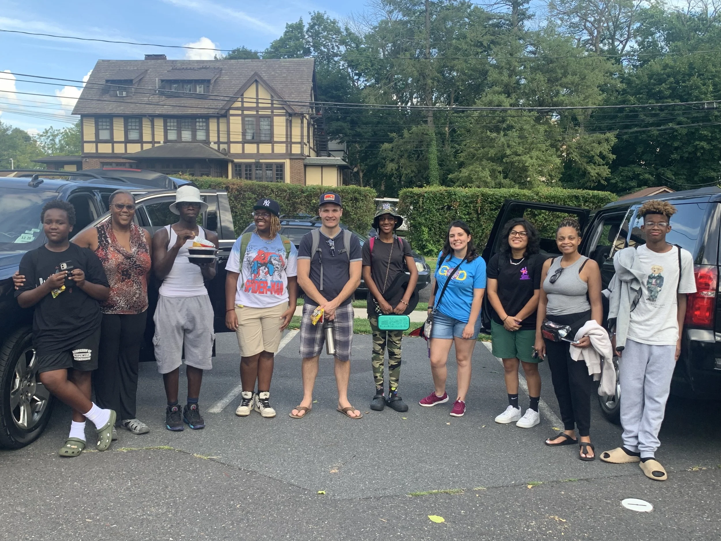 Group of eleven people standing in a parking lot in front of trees and parked cars, with a large yellow house with brown trim in the background.