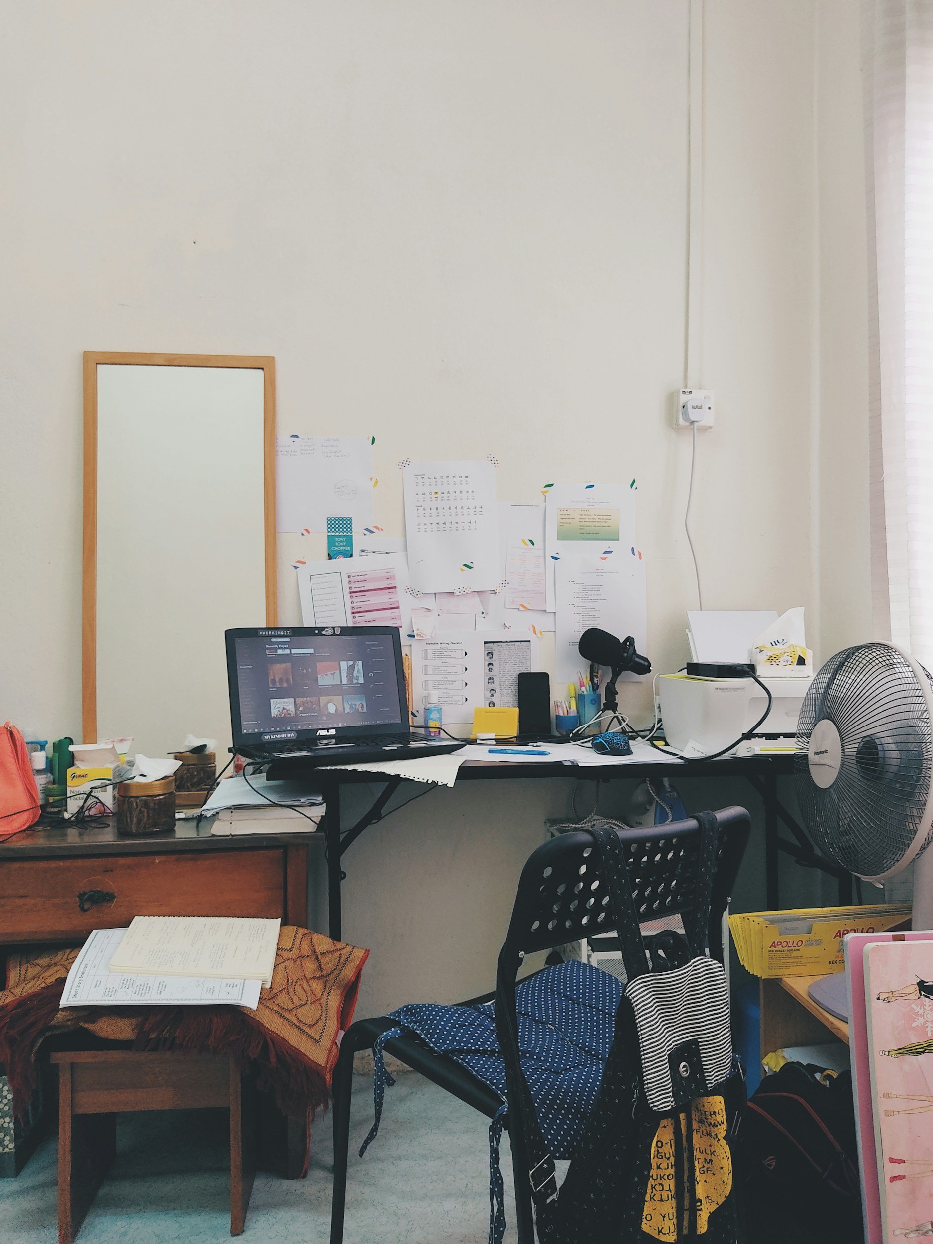 A cluttered desk in a home office with a laptop, papers, and office supplies. There is a chair with a backpack, a fan, a wooden side table with a box of tissues, and a corkboard with papers on the wall.