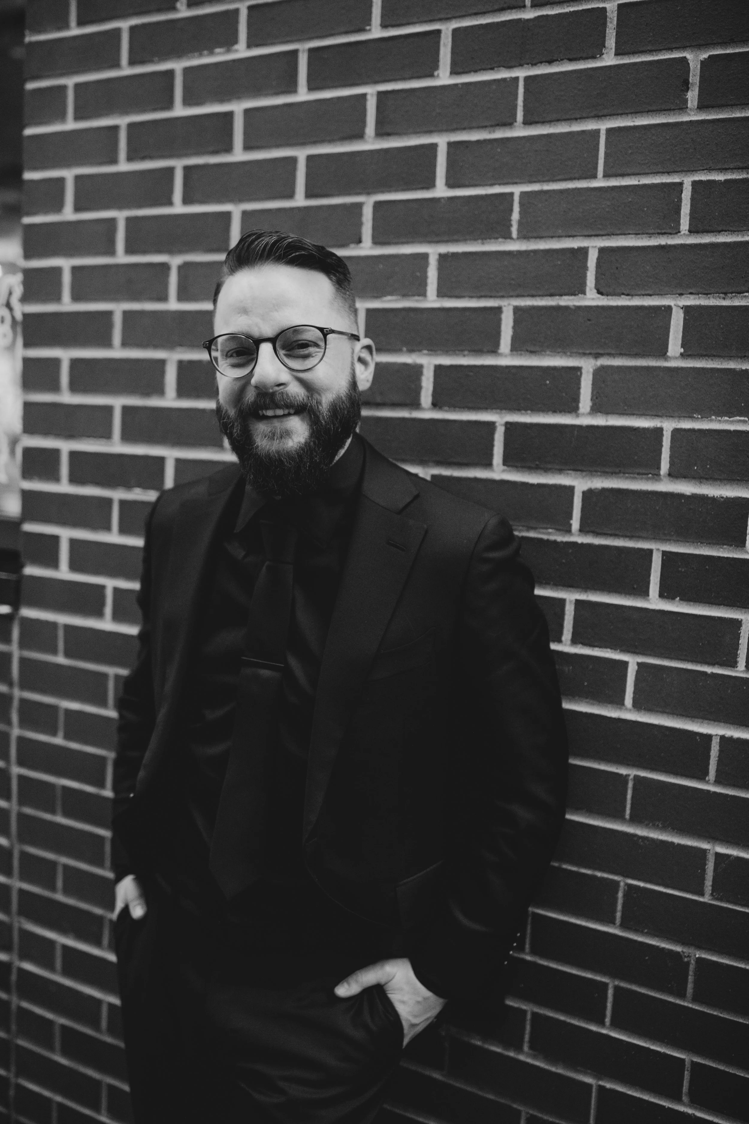 A man with glasses, a beard, and a smile, dressed in a black suit and tie, standing against a brick wall.