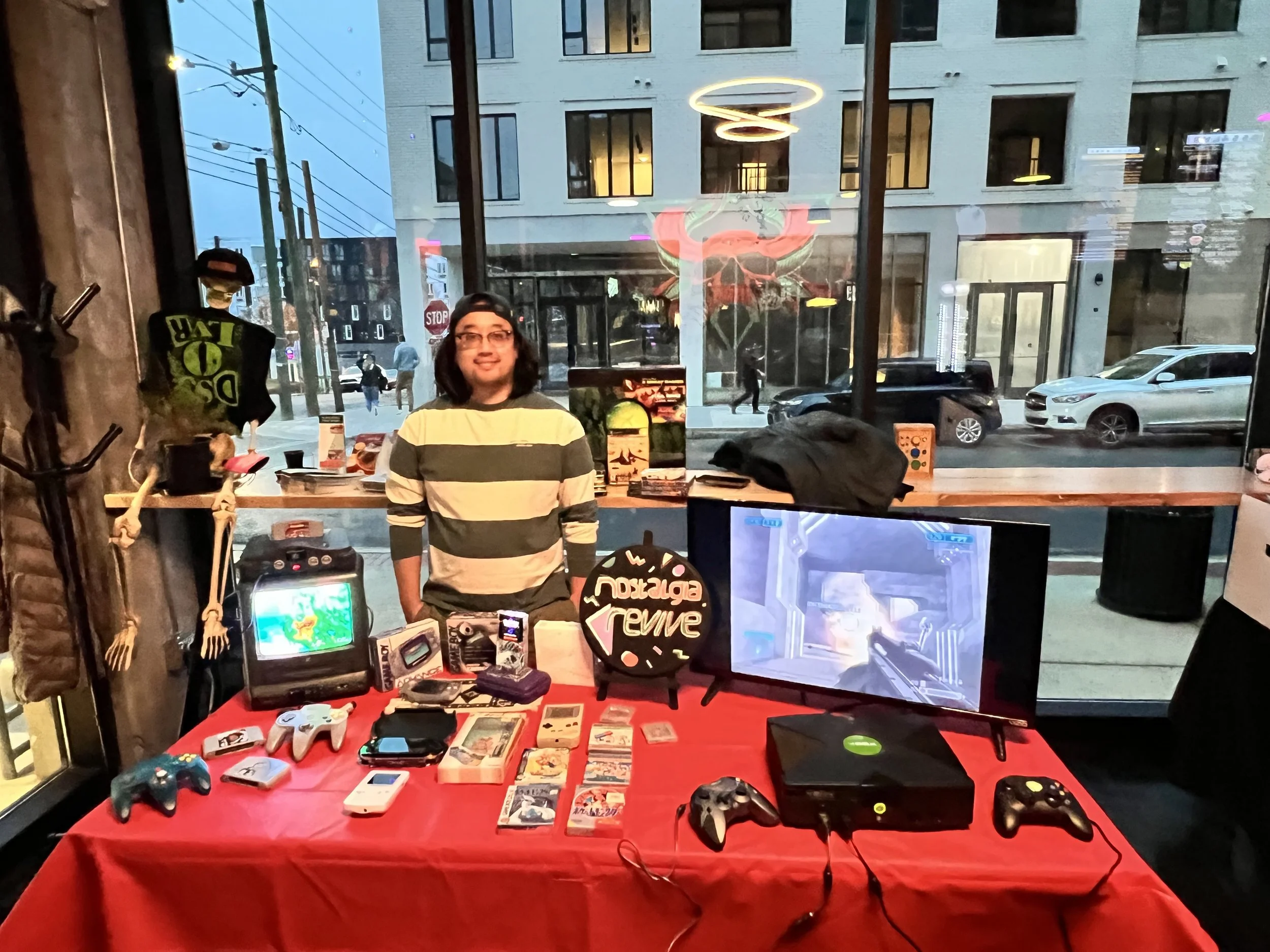 Young man standing behind a red table with retro gaming consoles, controllers, and video games on display inside a store with a large window showing a city street outside.