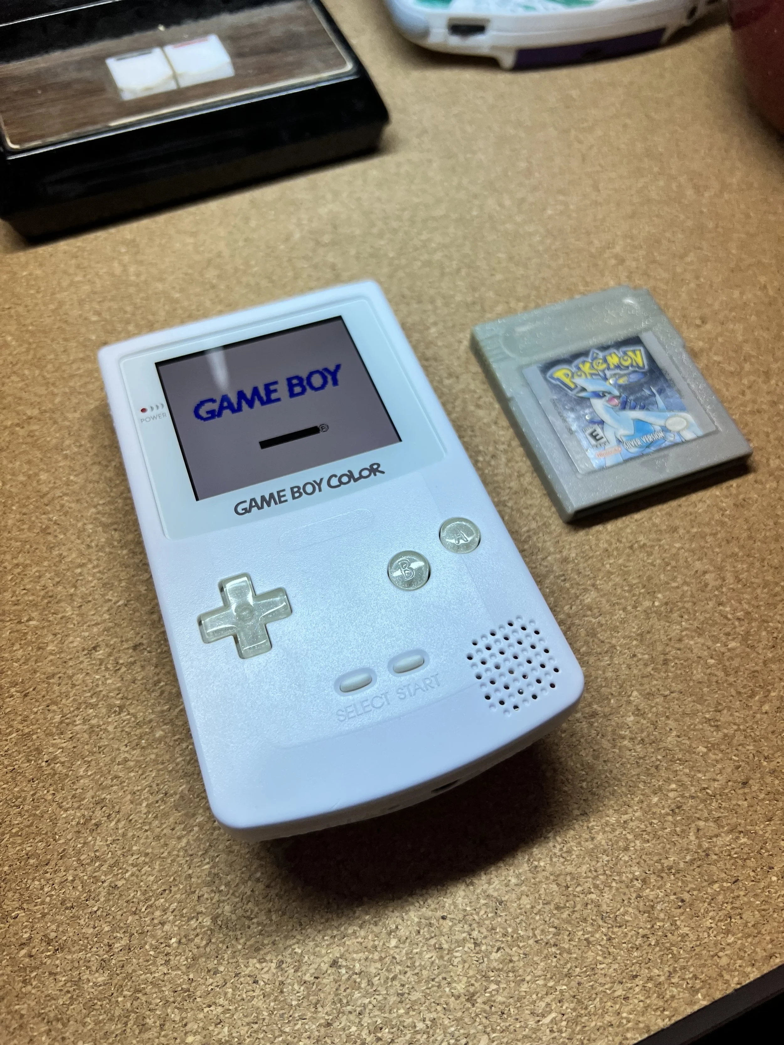 A white Nintendo Game Boy Color with a game menu on the screen that says 'GAME BOY,' next to a Pokémon game cartridge, on a wooden surface.