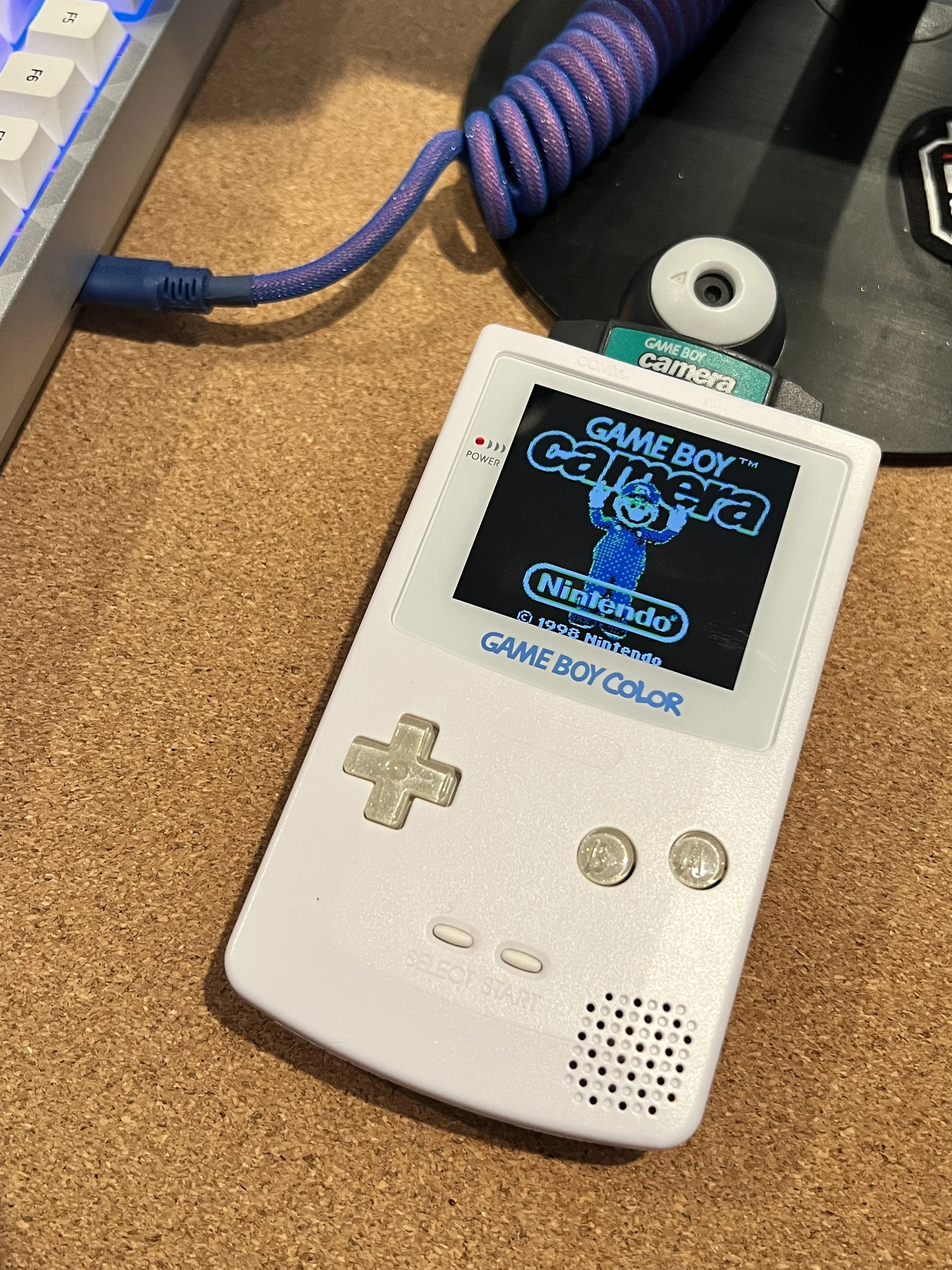 White Nintendo Game Boy Color displaying the title screen of the game 'Game Boy Camera' with a pixelated figure, on a wooden desk next to a purple coiled cable and a keyboard.