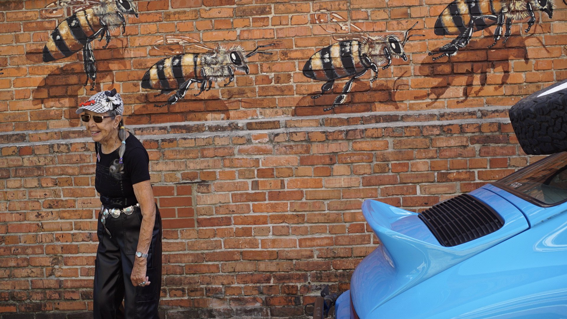An elderly woman with gray braids, wearing sunglasses, a black t-shirt, and black pants, walking past a brick wall featuring a mural of bees painted on it. There is a blue car partially visible on the right side of the image.