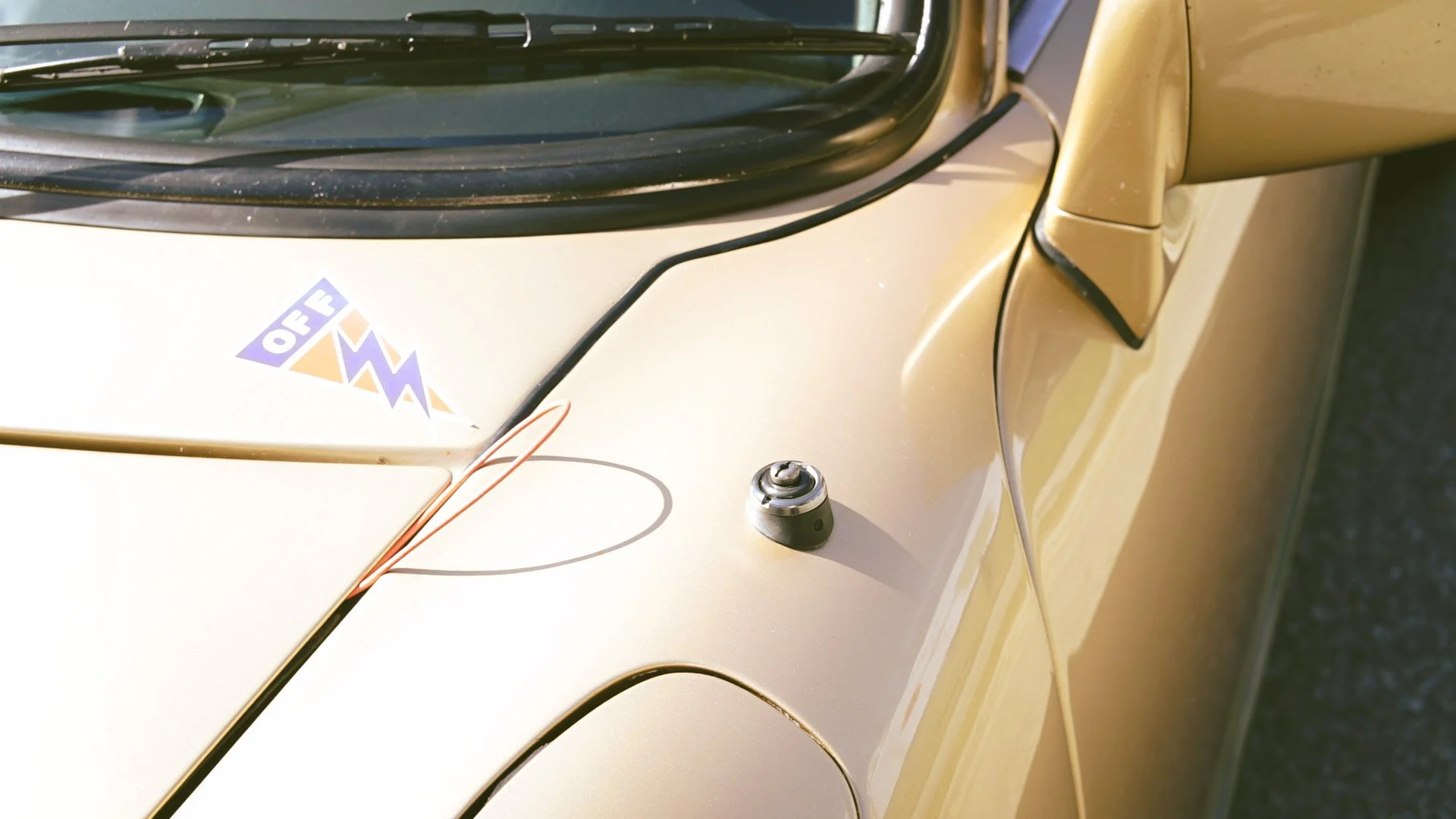 Close-up of a yellow car's hood with a black wire and an antenna base, and a sticker that reads 'OFF' with lightning bolt symbols.