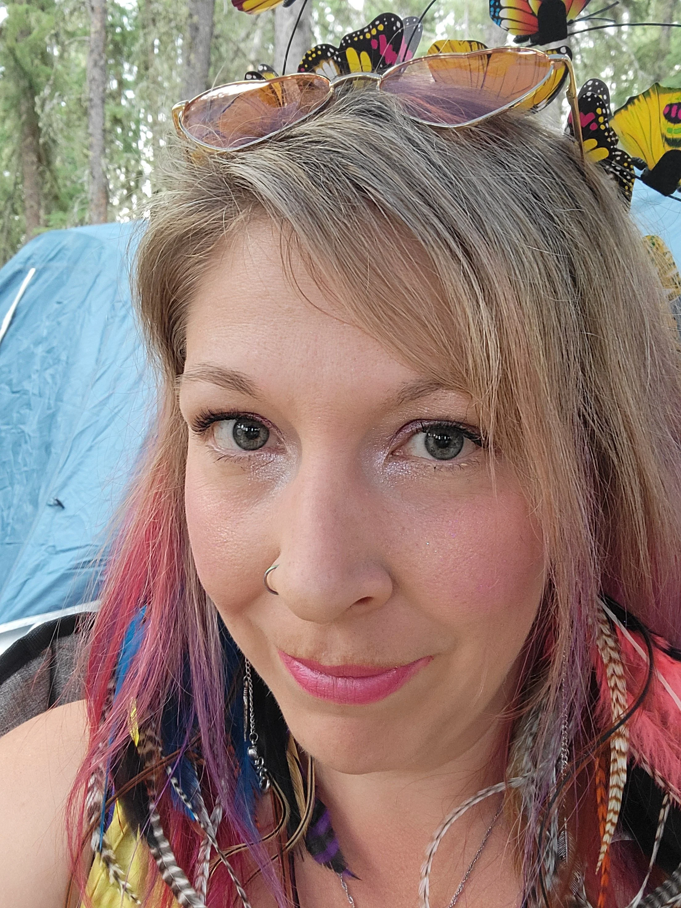 Picture of Shantay Karhut, owner of Celestial Spark. She has pinon feather earings and heart shaped glasses on top of her head. She is wearing light makeup with sparkly eyeshadow. Her hair is blonde with pink and purple tips.