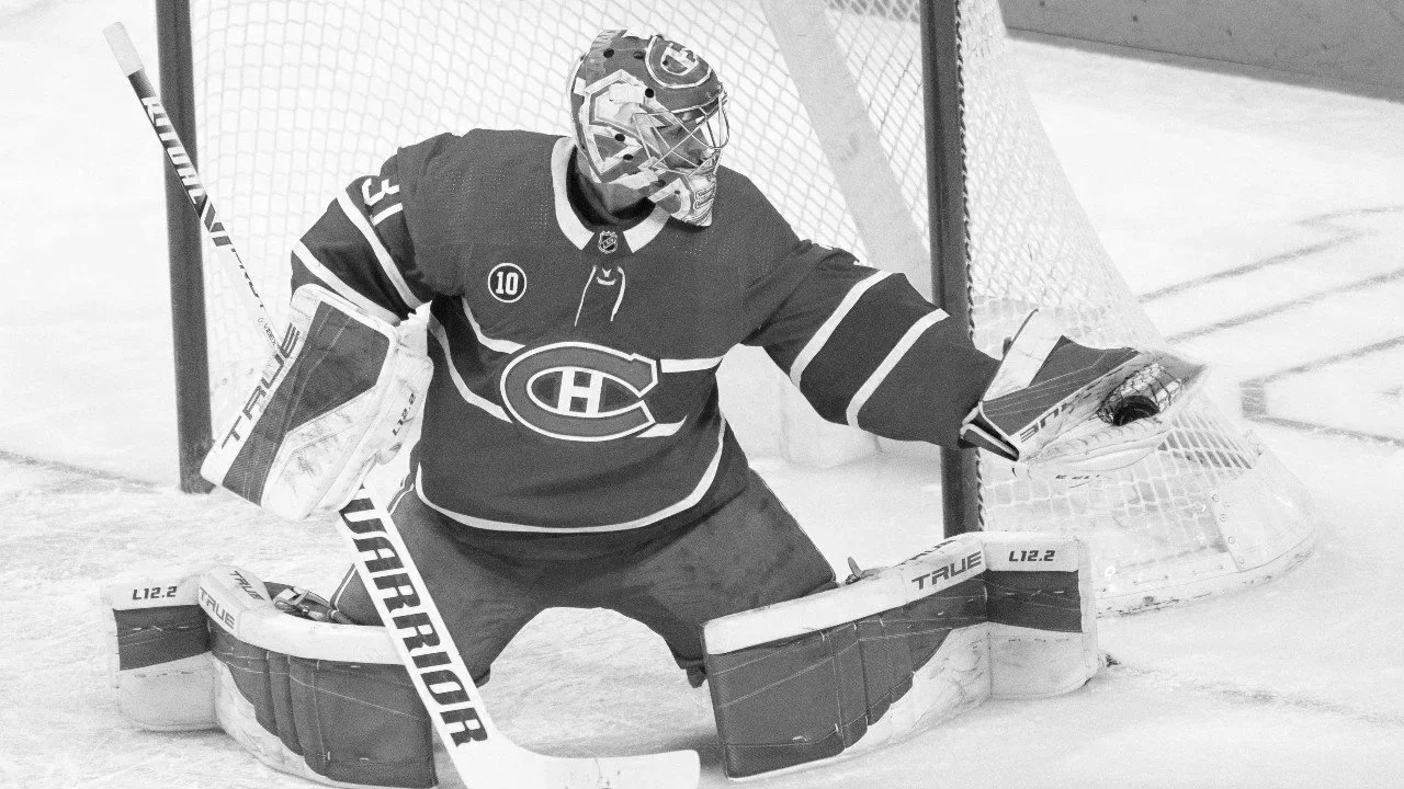 Hockey goalie in a uniform with a logo on the chest, wearing a helmet with a cage, standing in front of the net on the ice, prepared to block a shot.