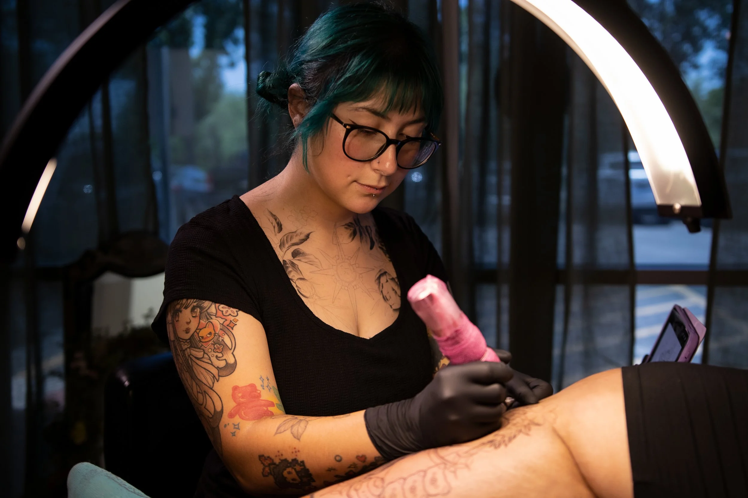 Victoria Tattoos artist working on a custom tattoo with machine wrapped in pink grip tape, floral neck tattoo visible, San Antonio tattoo studio.