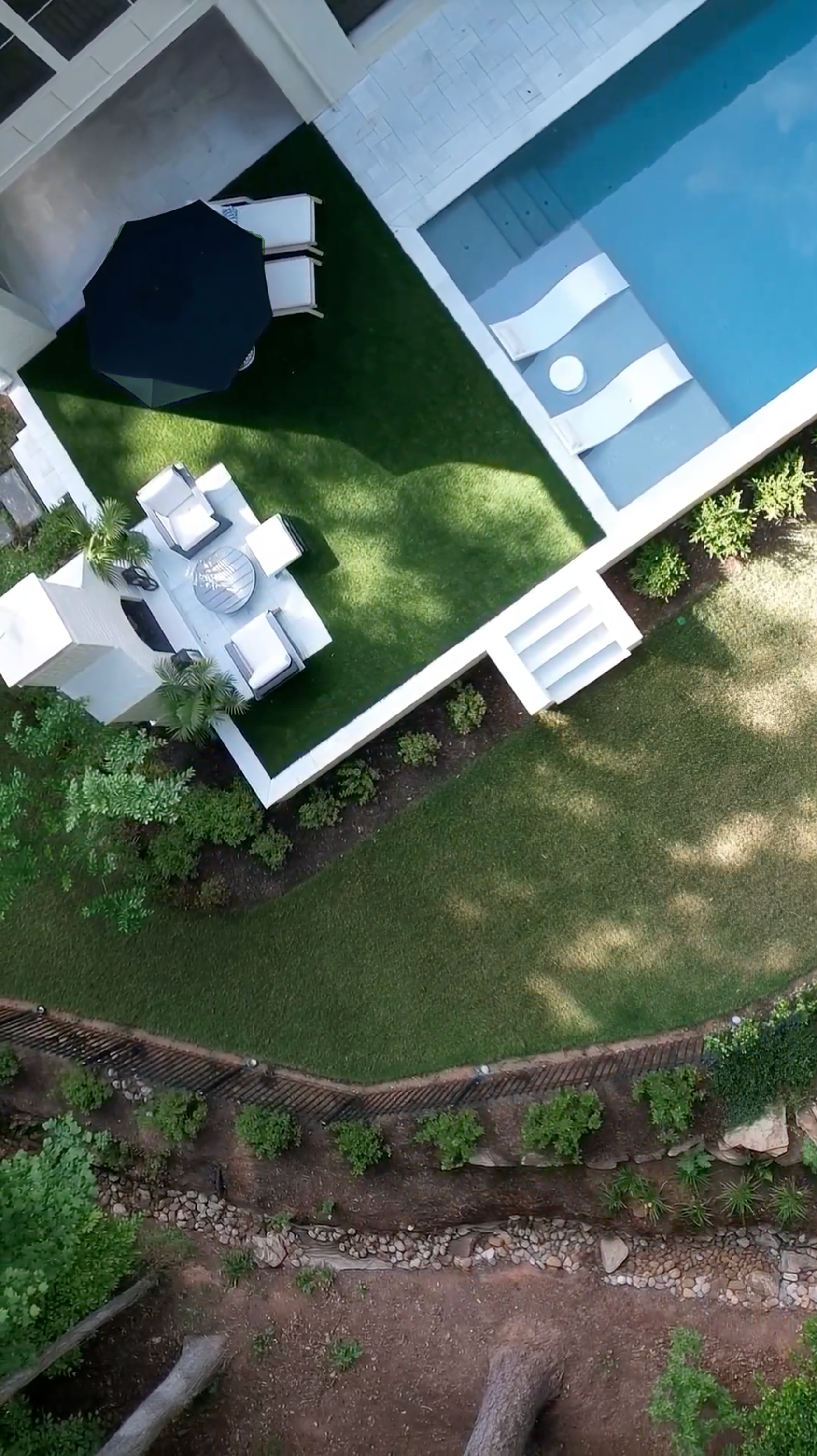 A landscaped terraced backyard with a green lawn, outdoor seating, outdoor fireplace, and a swimming pool, surrounded by plants and stormwater drainage featuring natural stones and custom fencing.
