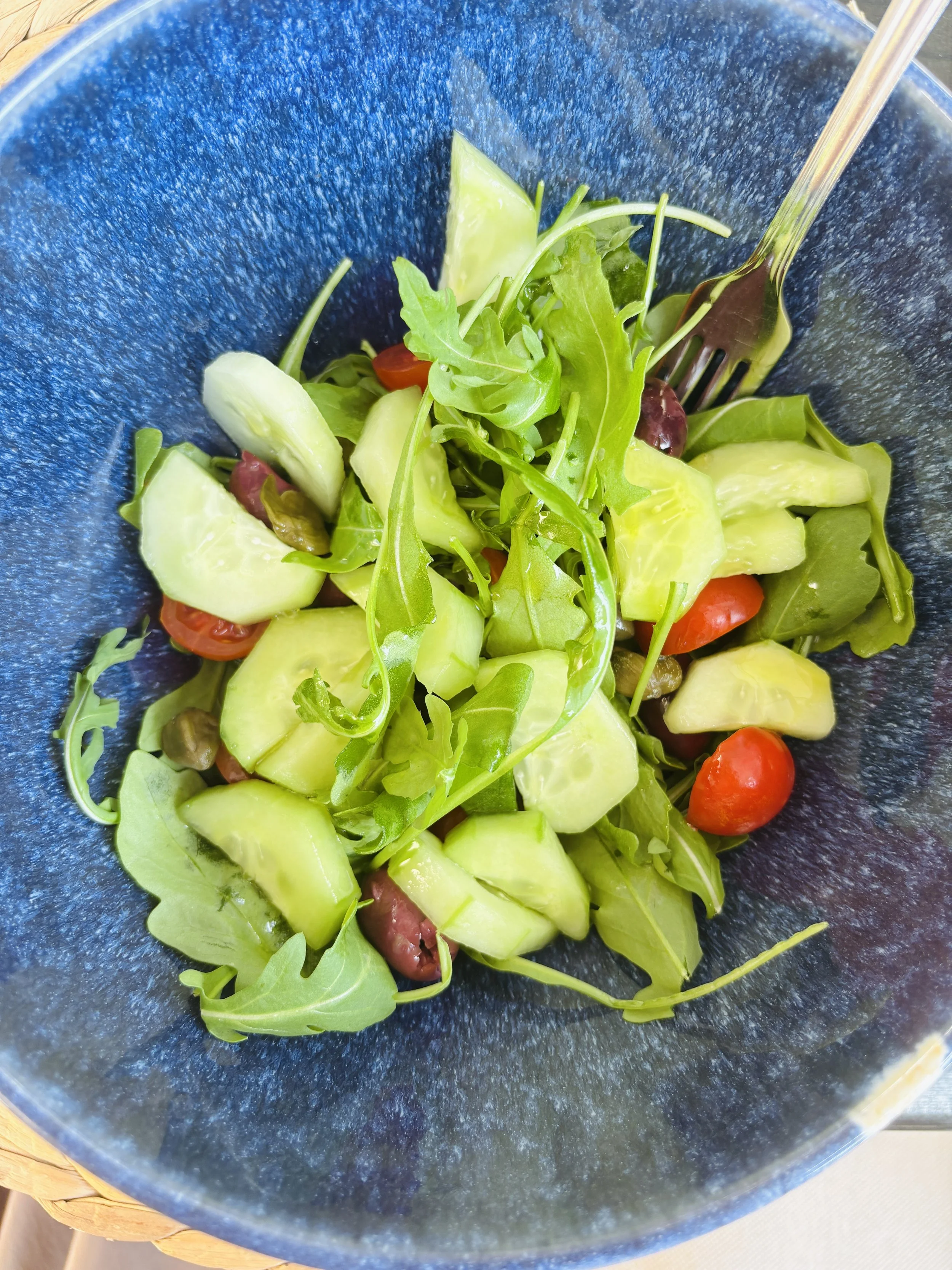 A green salad in a blue bowl containing arugula, cucumber, cherry tomatoes, green olives, and green bell peppers, with a fork resting on the rim.