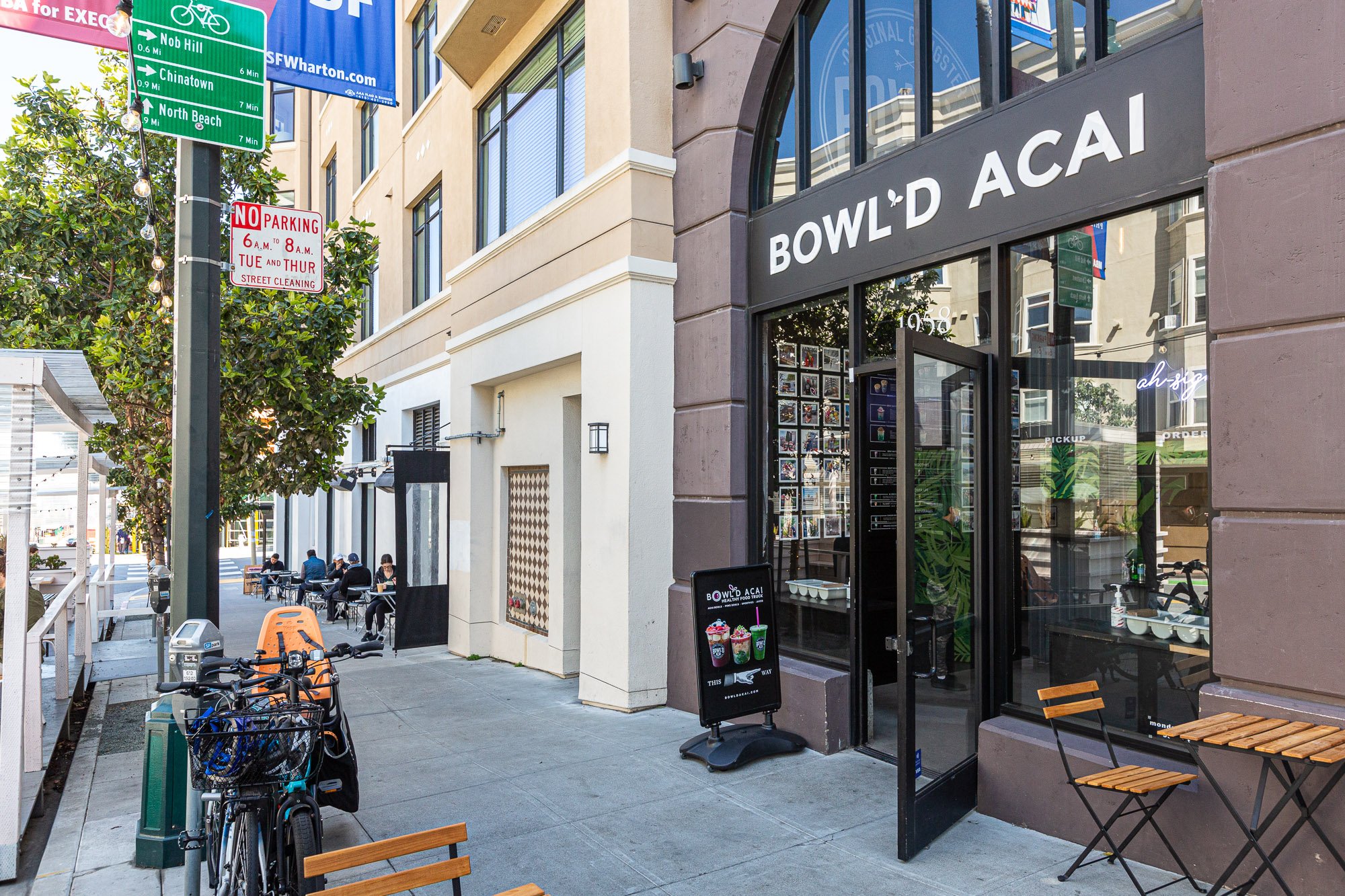 Street view of a restaurant named 'Bowld Acai' with outdoor seating, bicycles, and neighborhood signage including a no parking sign and street directions.