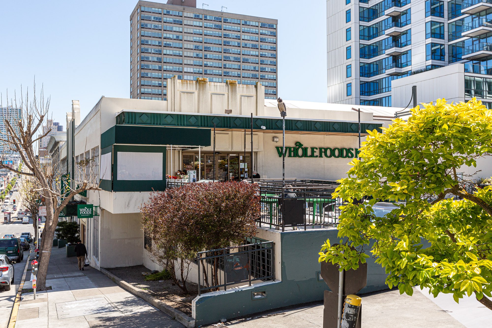 Exterior of Whole Foods Market building in an urban area with sidewalks, trees, and tall modern buildings in the background.